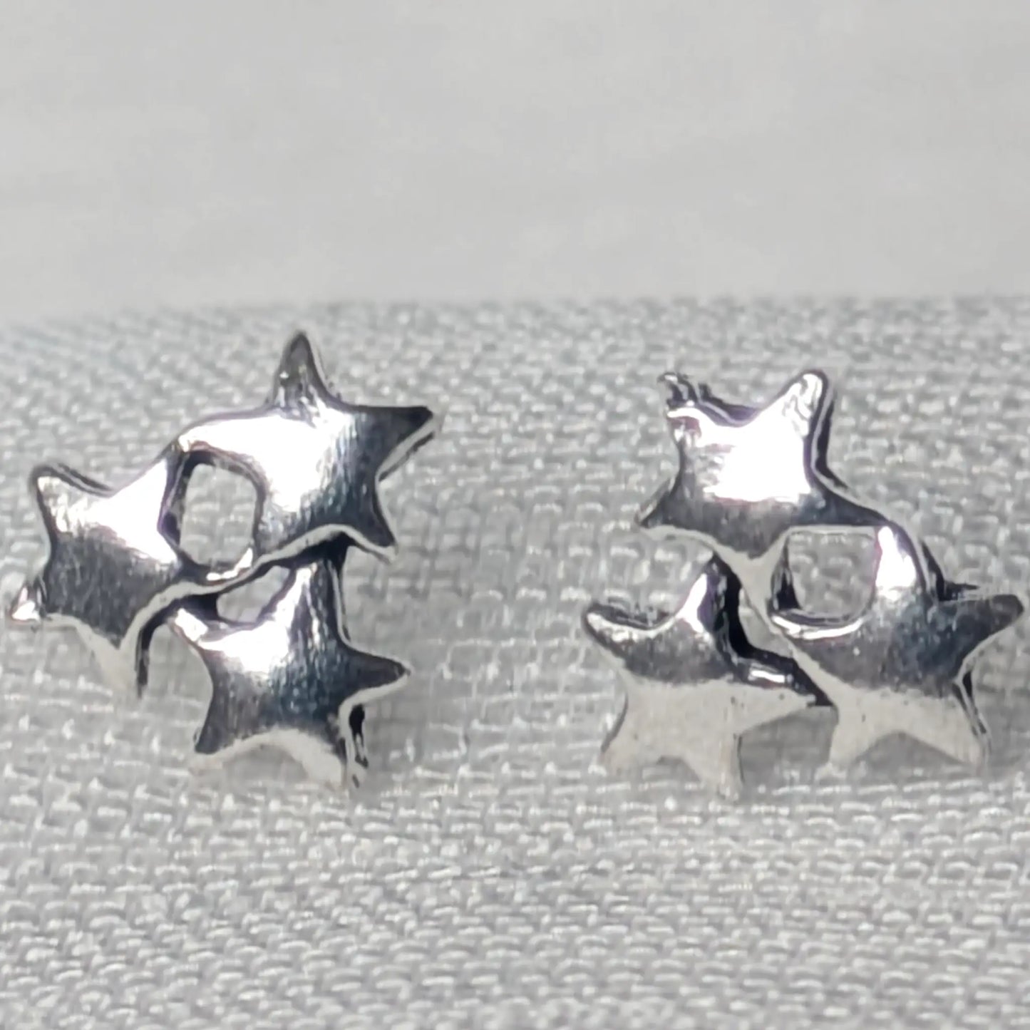 Detaillierte Nahaufnahme von zwei glänzenden Dreifach-Stern-Ohrsteckern aus 925 Sterling Silber auf einer hellen Gewebestruktur