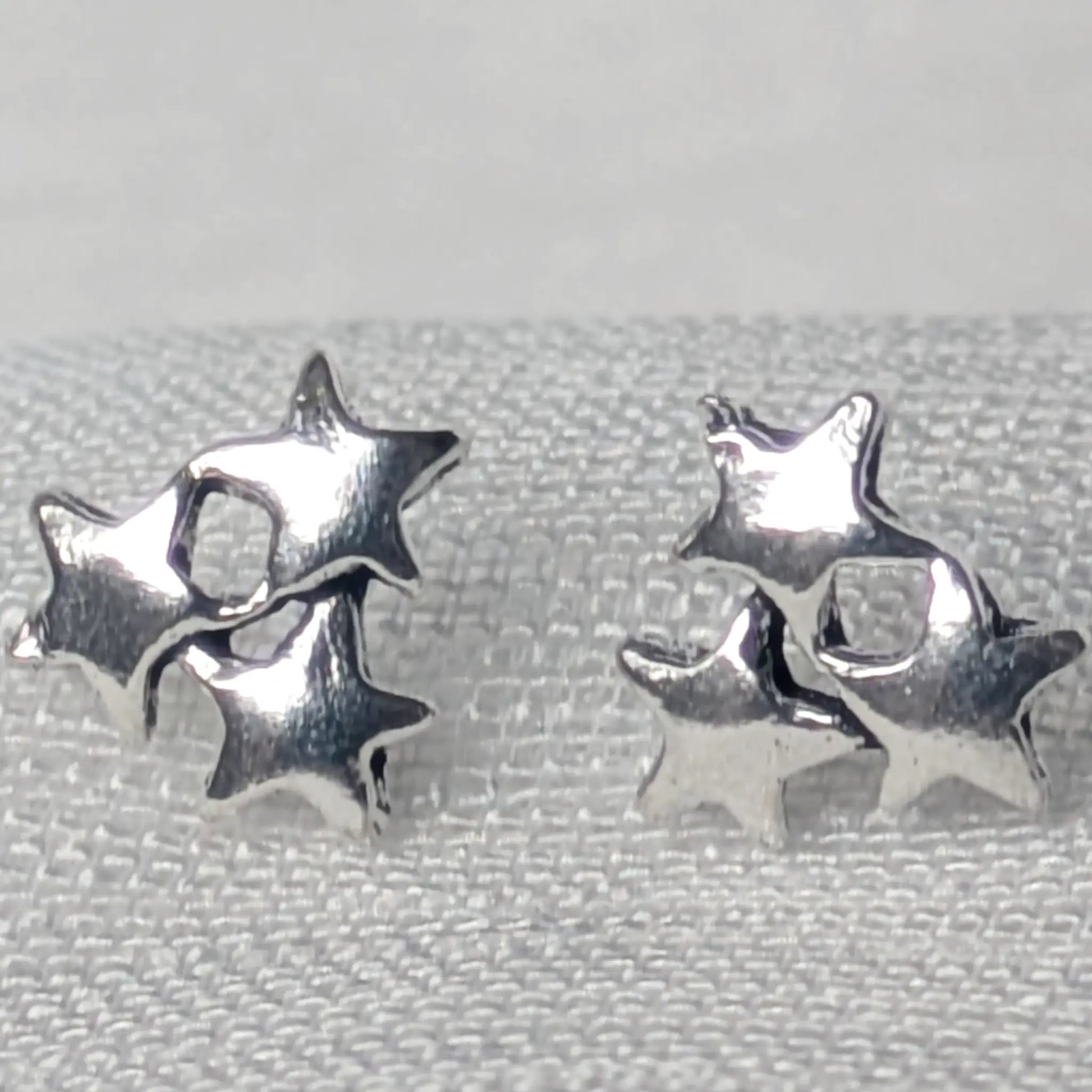 Detaillierte Nahaufnahme von zwei glänzenden Dreifach-Stern-Ohrsteckern aus 925 Sterling Silber auf einer hellen Gewebestruktur