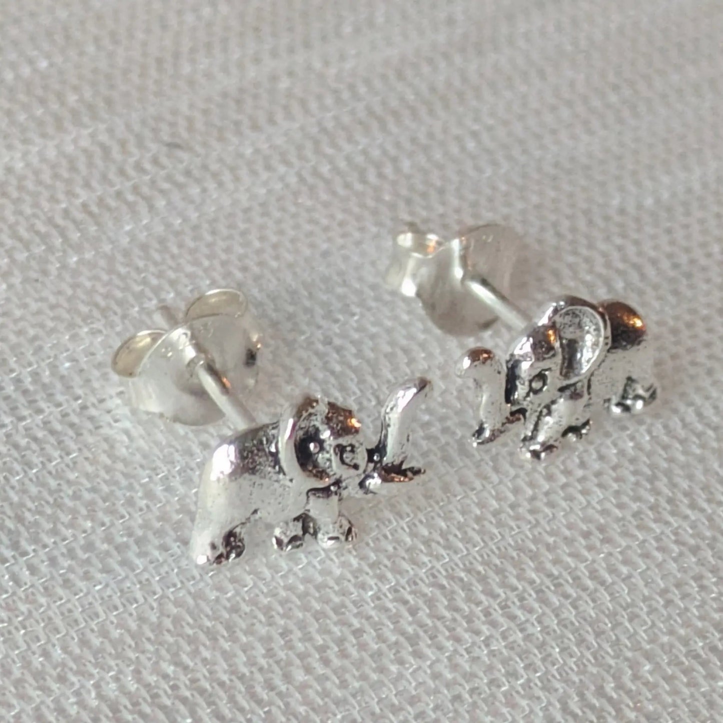 Zwei filigrane Elefanten-Schmuckstücke als Ohrstecker auf einem leicht strukturierten, weißen Stoff, die die Details hervorheben.