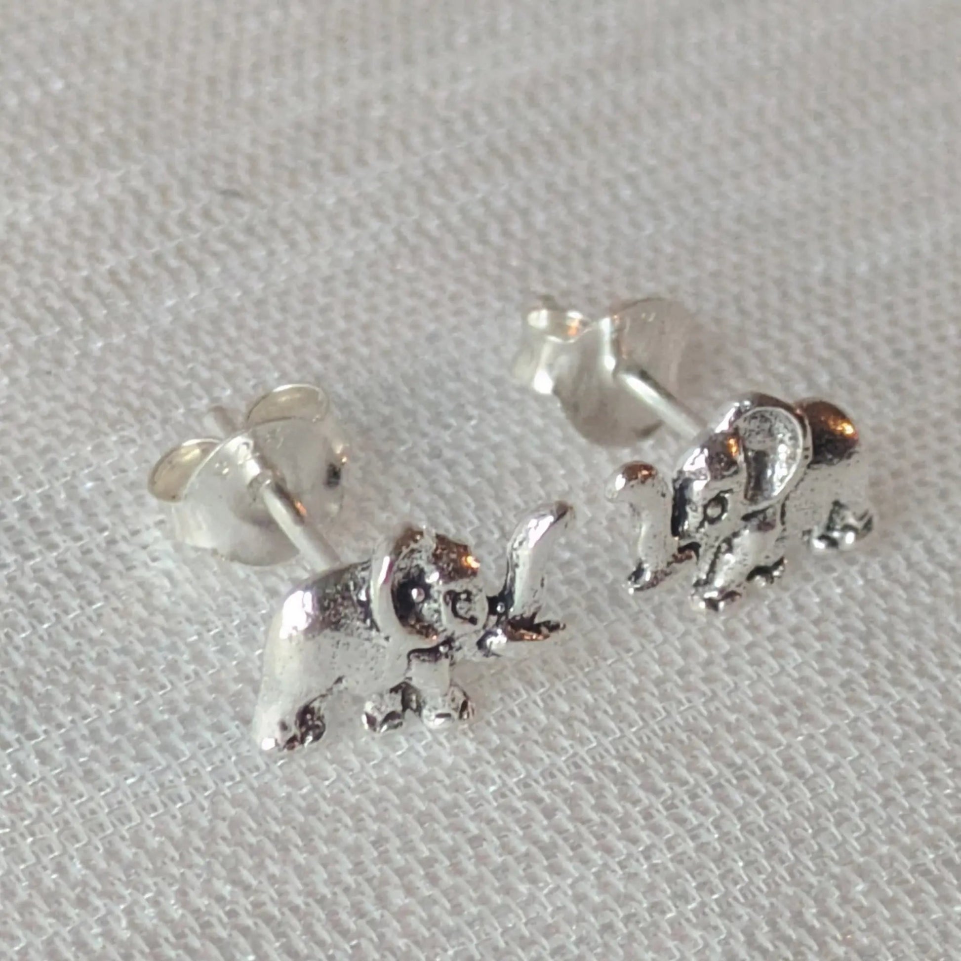 Zwei filigrane Elefanten-Schmuckstücke als Ohrstecker auf einem leicht strukturierten, weißen Stoff, die die Details hervorheben.