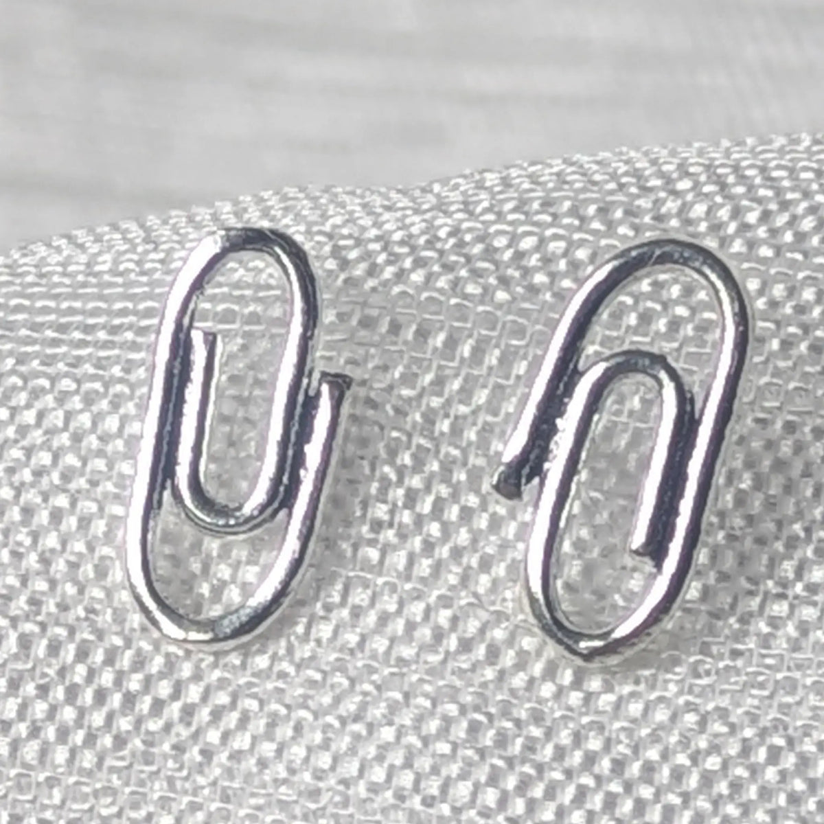  Detailaufnahme von zwei glänzenden 925 Silber Ohrsteckern im minimalistischen Büroklammer-Design auf hellem Stoff.