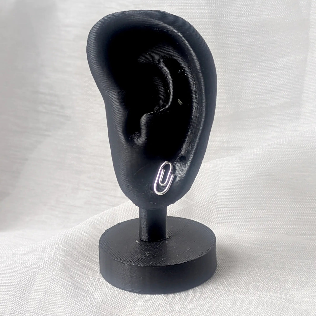 Ein silberner Büroklammer-Ohrstecker dezent platziert an einem schwarzen 3D-Ohr-Display zur Veranschaulichung der Proportionen.
