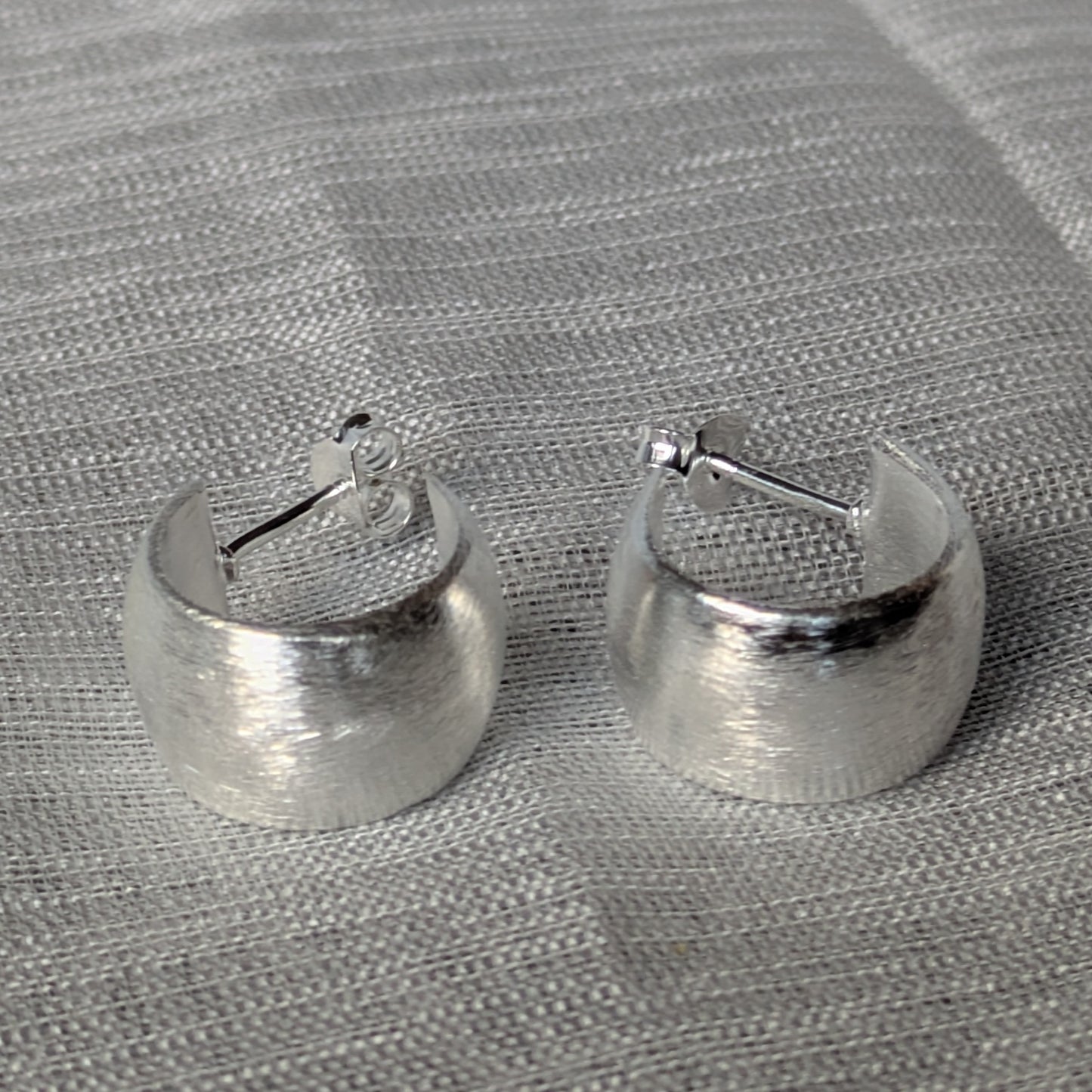 Breite Creolen aus 925 Silber mit gebürsteter Oberfläche auf grauem Stoffhintergrund.
