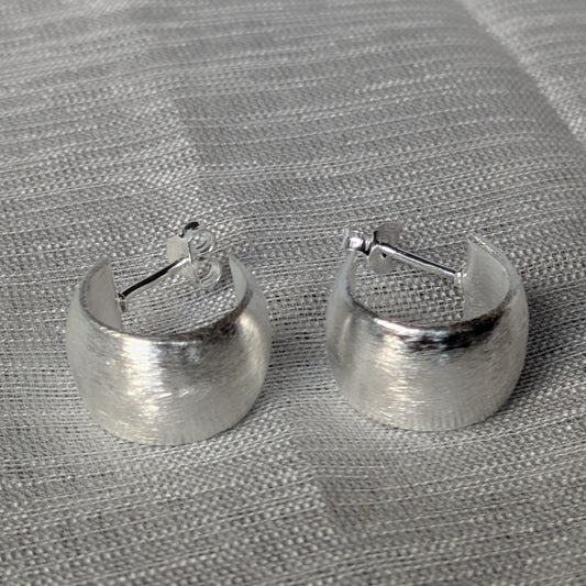 Breite Creolen aus 925 Silber mit gebürsteter Oberfläche auf grauem Stoffhintergrund.