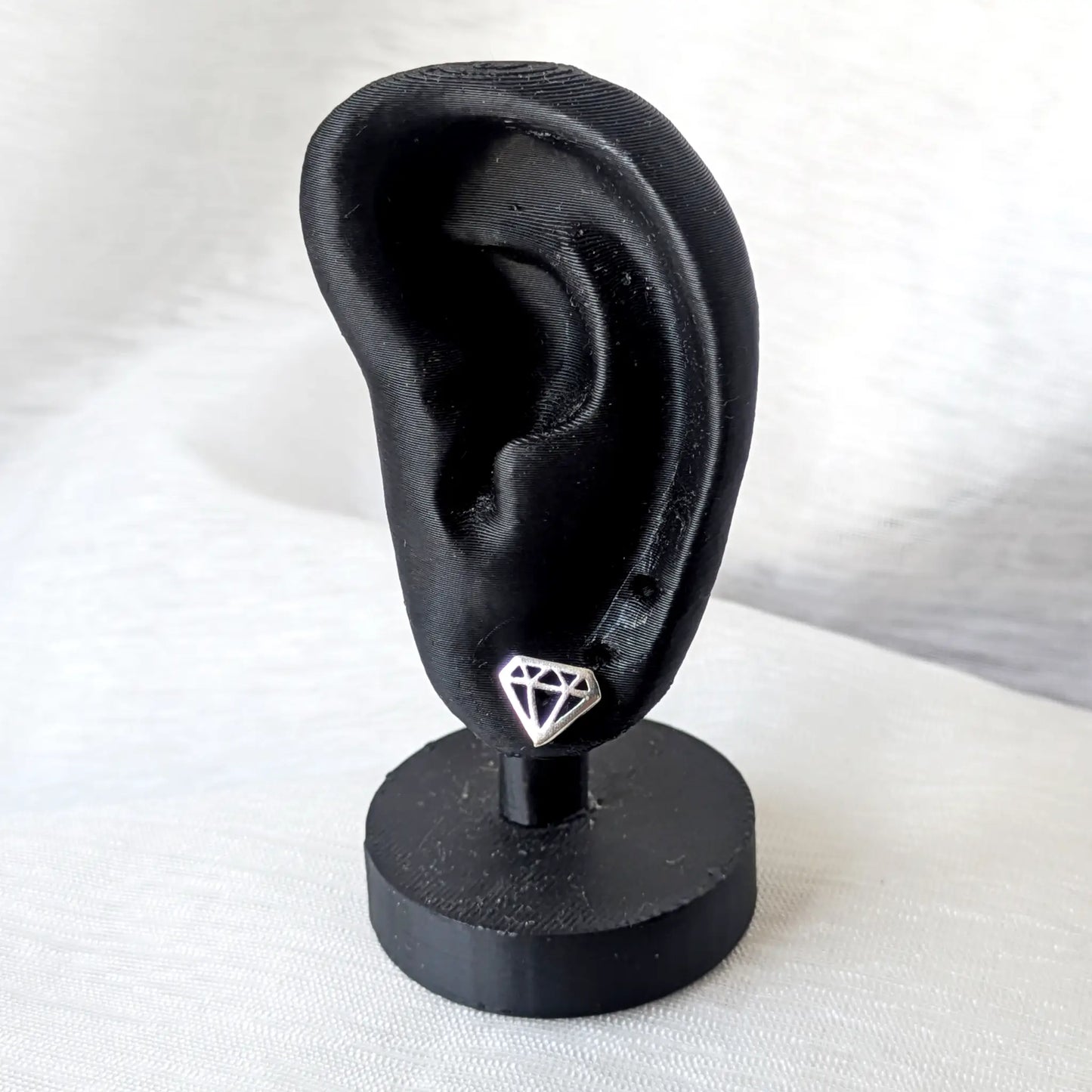 Ein minimalistischer Diamanten-Form Ohrstecker aus Silber getragen an einem mattschwarzen Deko-Ohr zur Größenansicht.