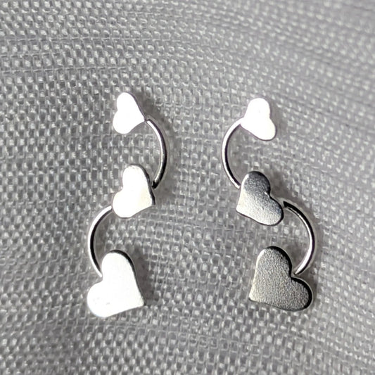 Silberne Herz-Ohrstecker mit drei Herzen in Reihe auf weißem Stoffhintergrund.