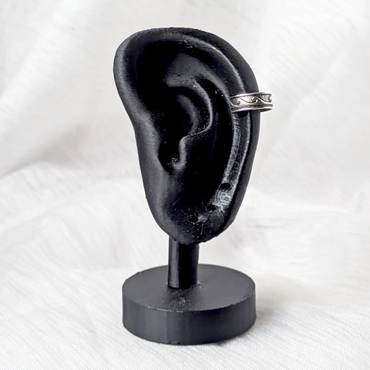 Silberner Ear Cuff im Wellen-Design, festgeklemmt am oberen Knorpel eines schwarzen 3D-Ohr-Displays.