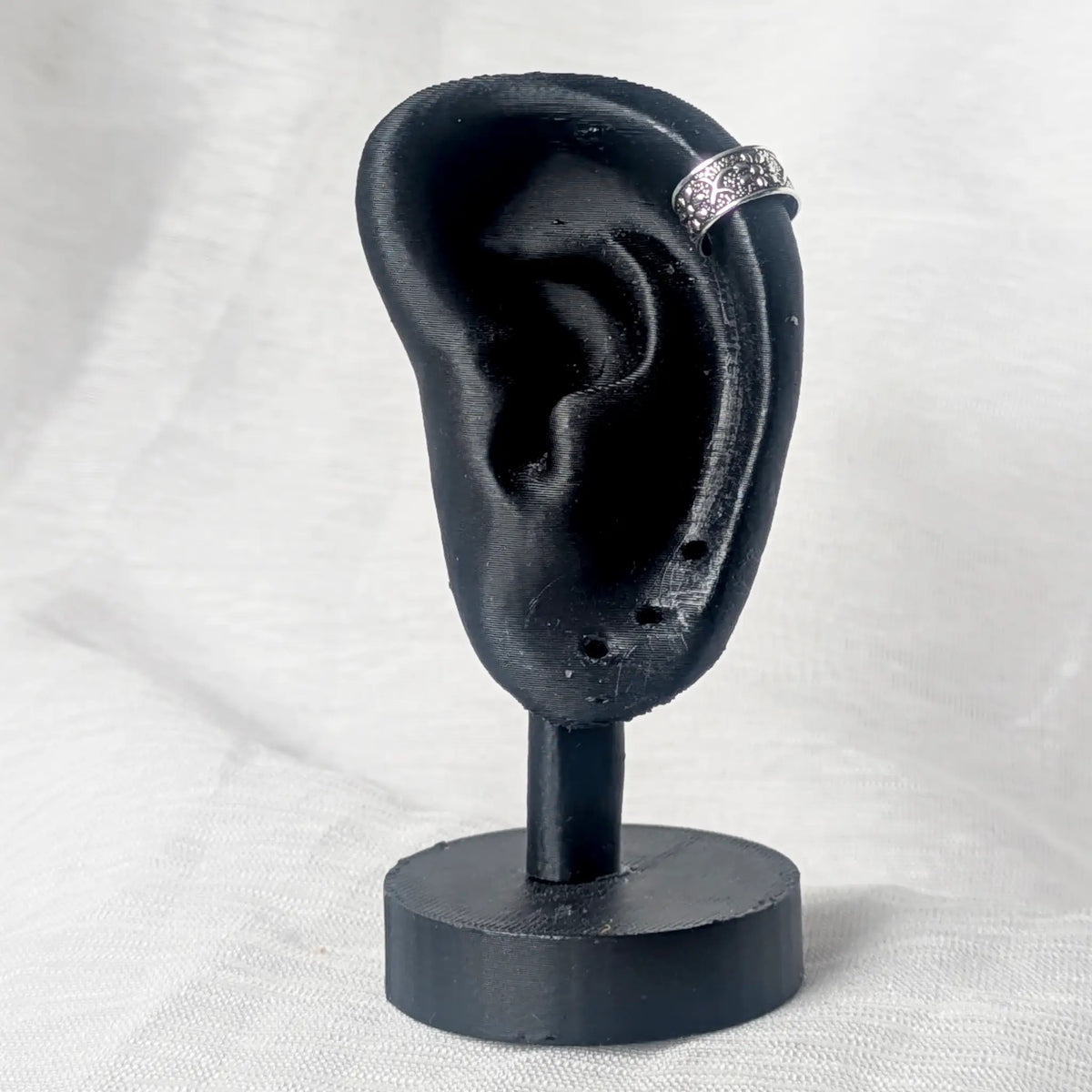Silberner Earcuff mit Blütenmuster, getragen an der oberen Knorpelkante (Helix) eines schwarzen Modell-Ohrs zur Demonstration.