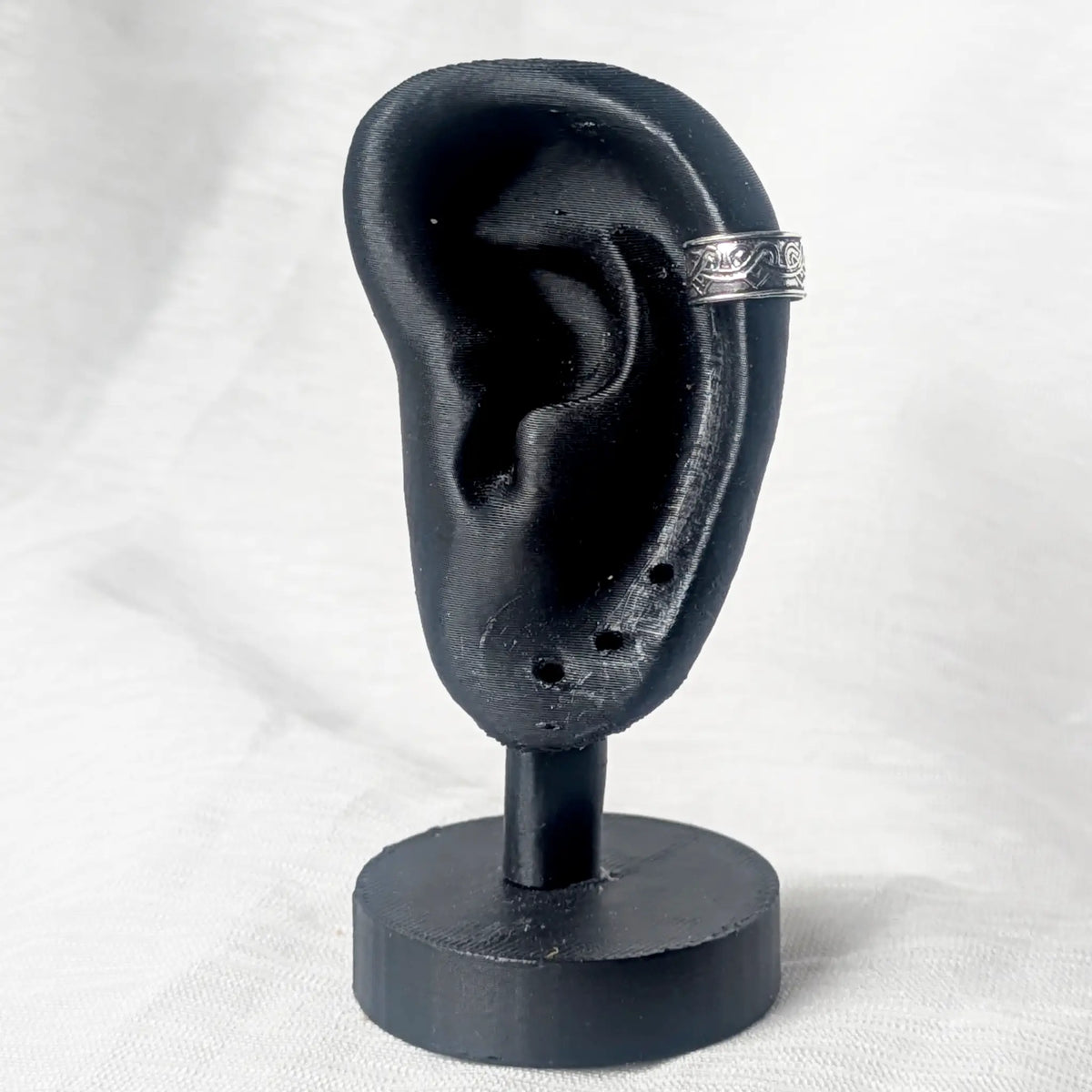 Silberner Earcuff mit Herz-Muster, platziert an der oberen Knorpelkante eines schwarzen Modell-Ohrs zur Demonstration der Passform.
