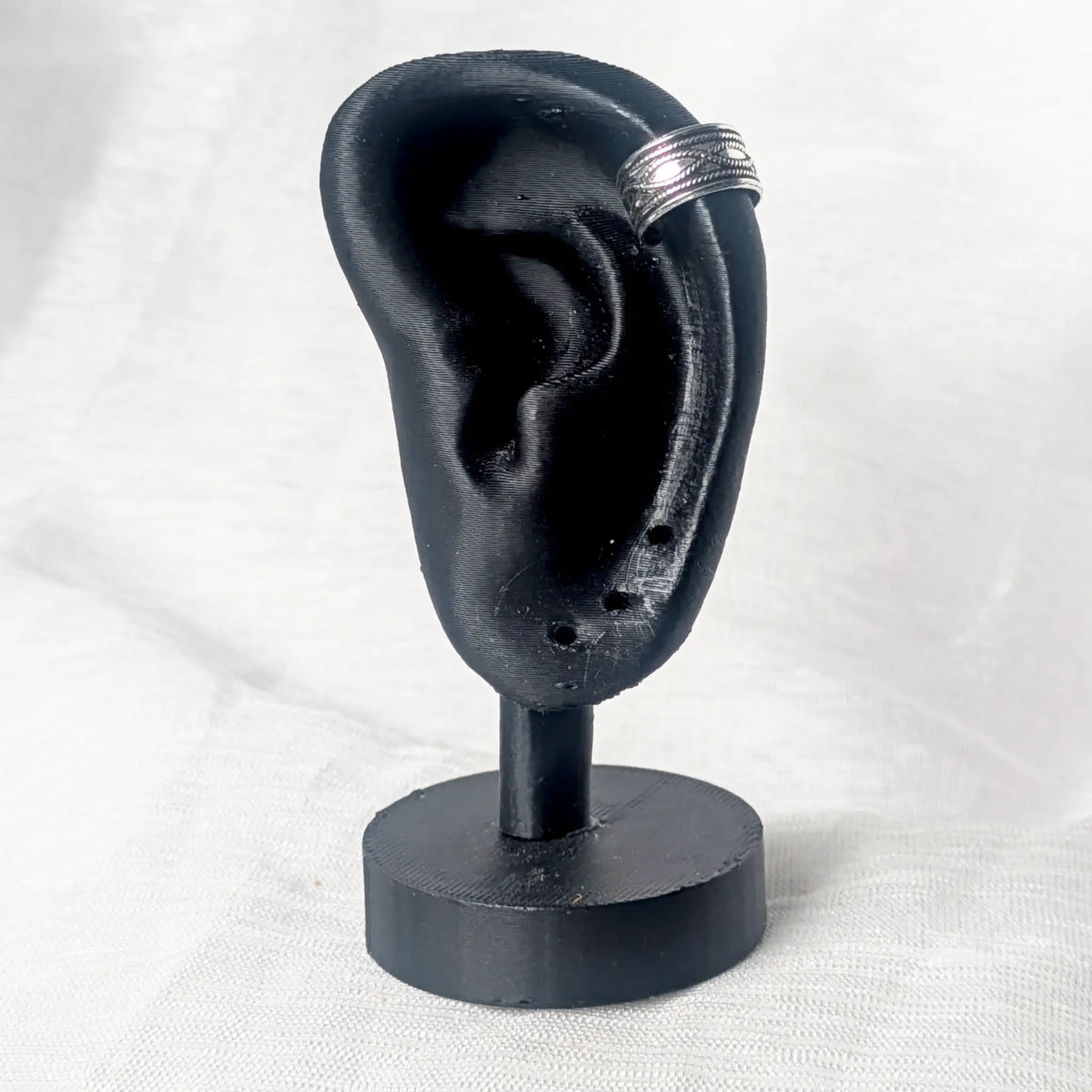 Silberner Earcuff mit Kordel-Design, getragen an der Helix-Position eines schwarzen Modell-Ohrs zur Veranschaulichung der Passform.
