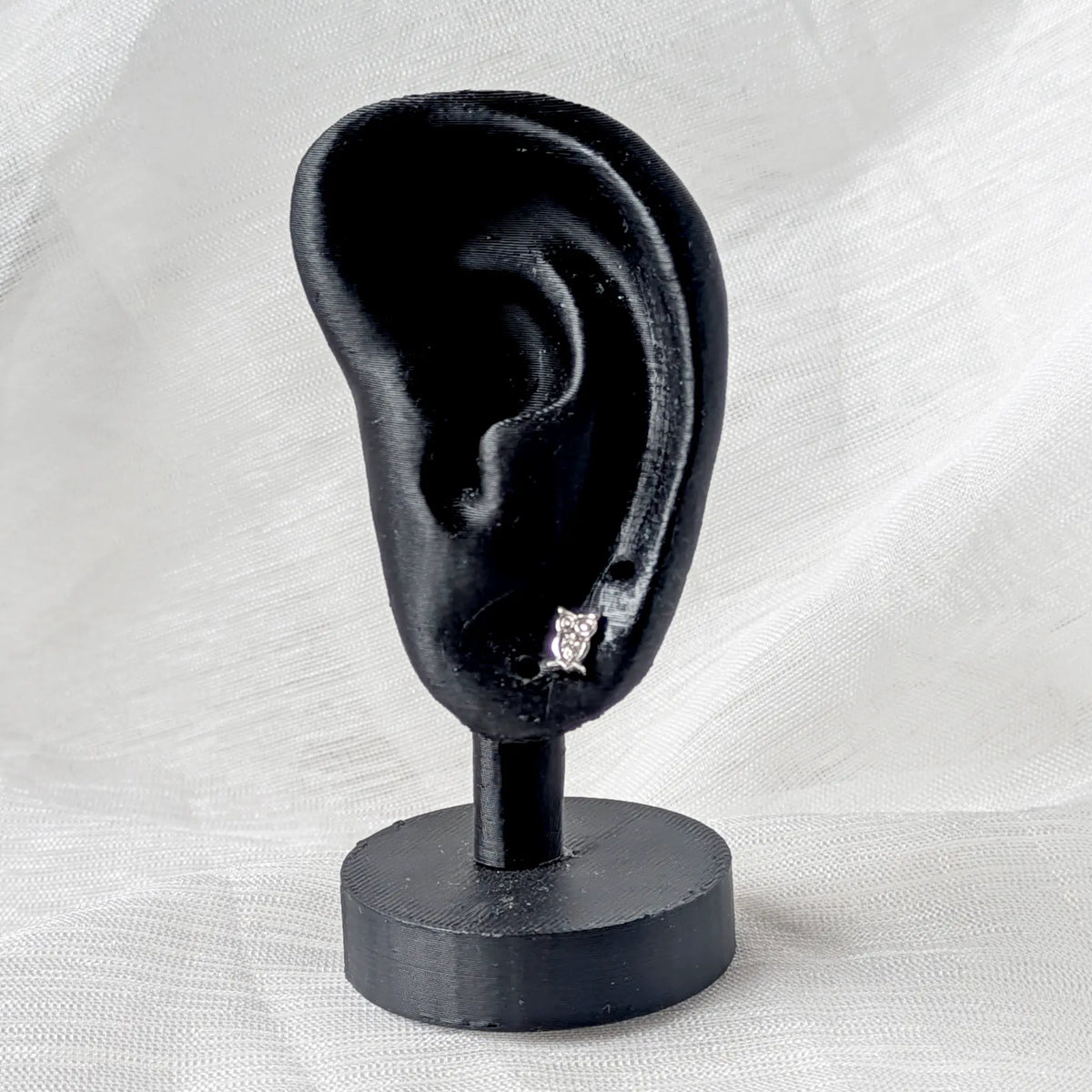 Ein kleiner Eulen-Silberohrstecker getragen an einem schwarzen 3D-Ohr-Modell zur Veranschaulichung der dezenten Größe und Passform.