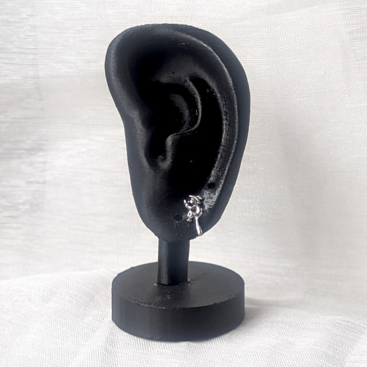 Ein silberner Hexe-auf-Besen Ohrstecker platziert an einem schwarzen 3D-Ohr-Display zur Veranschaulichung der Größe.