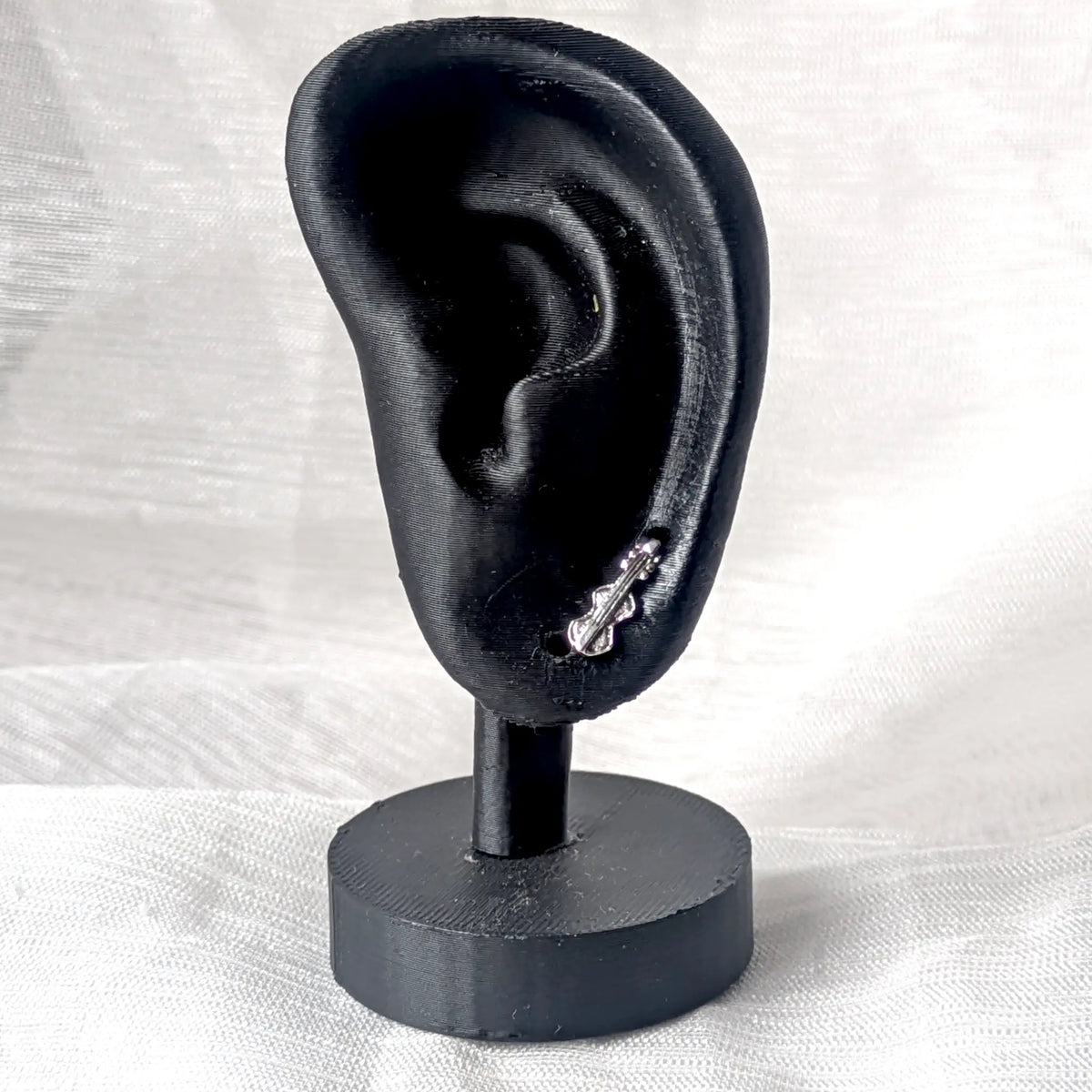 Ein silberner Geigen-Ohrstecker, angebracht an einem schwarzen 3D-Ohr-Display zur Anschaulichkeit der Proportionen am Ohrläppchen.