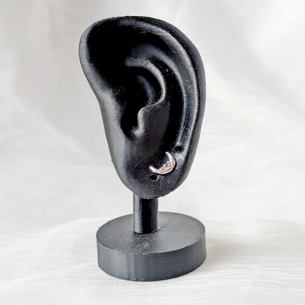 Ein kleiner silberner Halbmond-Ohrstecker präsentiert an einem schwarzen 3D-Ohr-Display, um die dezente Passform am Ohrläppchen zu zeigen.

