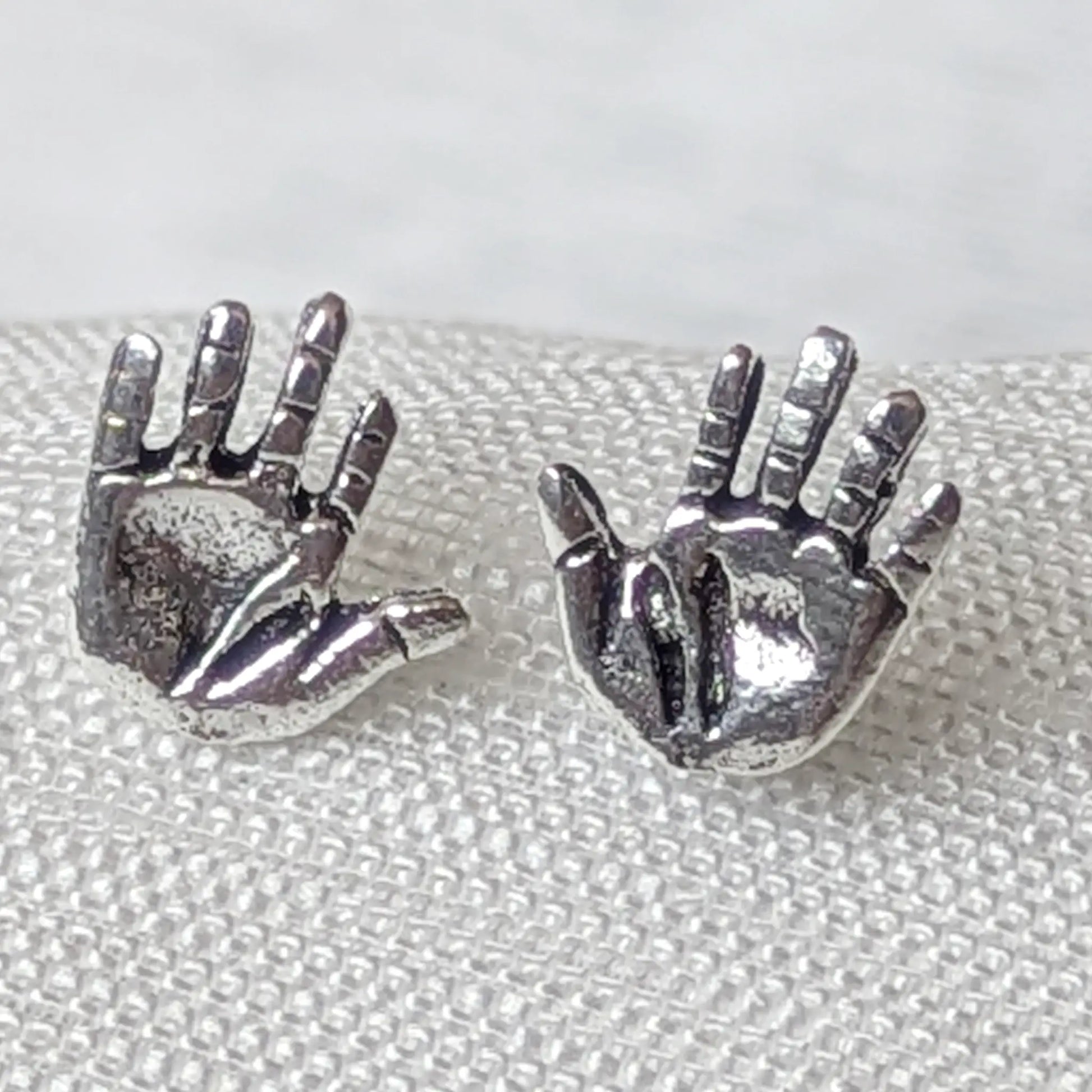 `Nahaufnahme von zwei detailreichen Ohrsteckern in Hand-Form aus geschwärztem 925 Sterlingsilber auf strukturiertem Stoff.