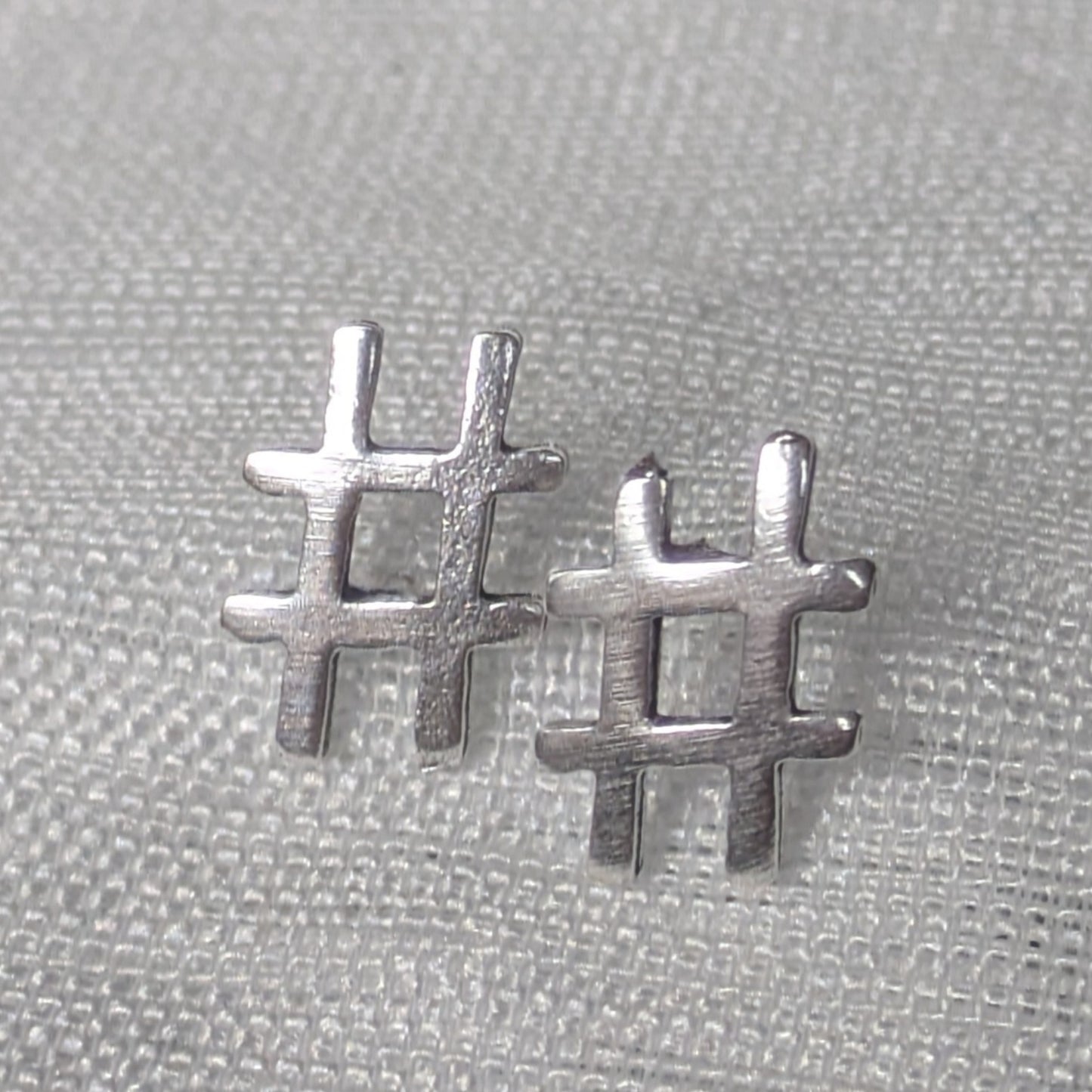 Frontalansicht der silbernen Hashtag-Ohrstecker mit mattierter Oberfläche auf hellem Stoff