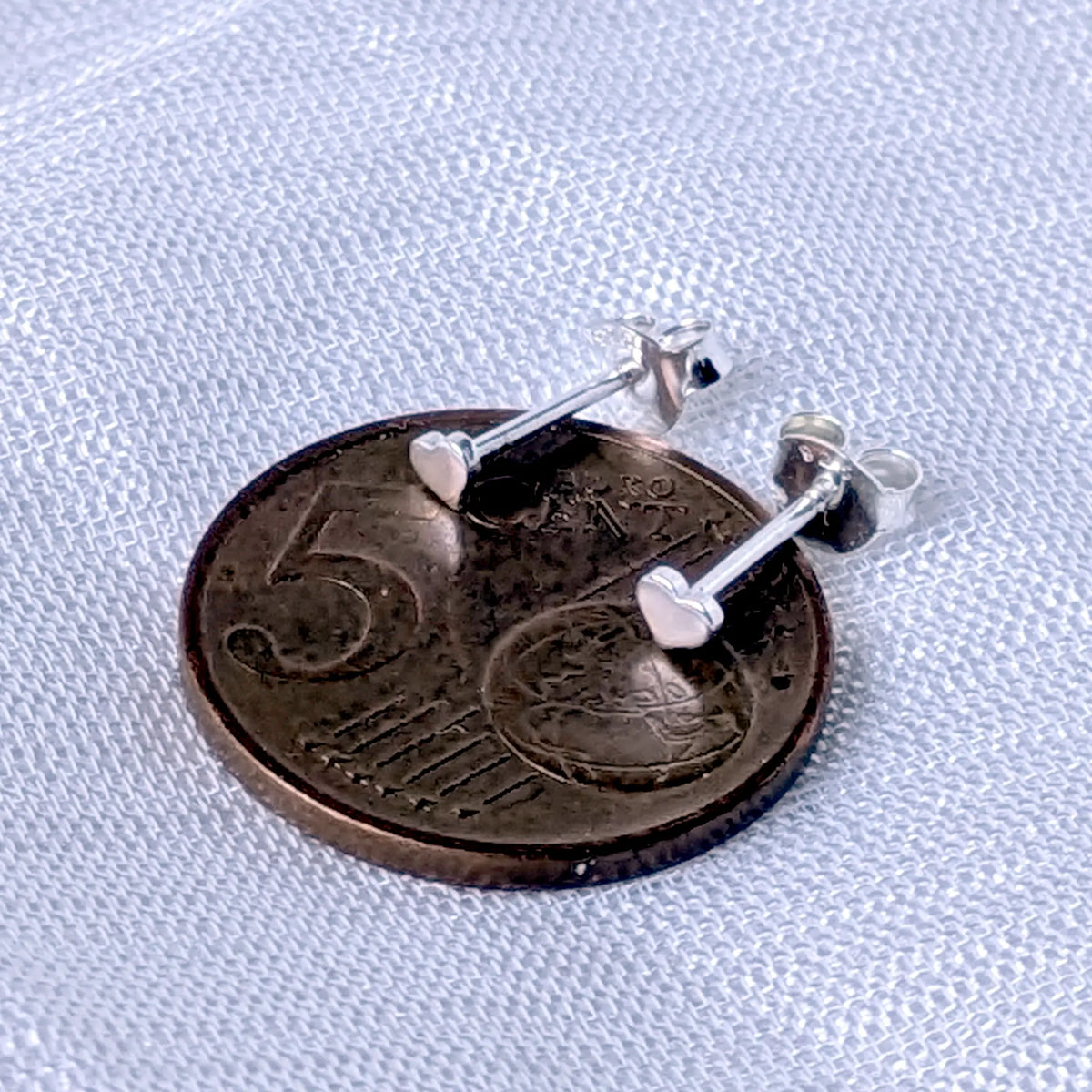 Kleine Herz-Silberohrstecker, direkt auf einer 5-Cent-Münze platziert, um die zierlichen Größen von 3mm zu veranschaulichen.