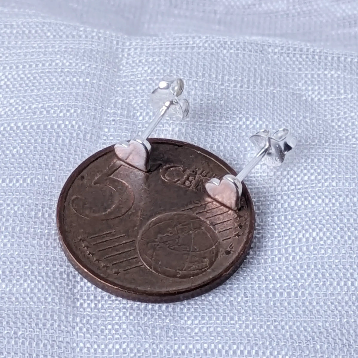 Kleine Herz-Silberohrstecker, direkt auf einer 5-Cent-Münze platziert, um die zierlichen Größen von 5mm zu veranschaulichen.
