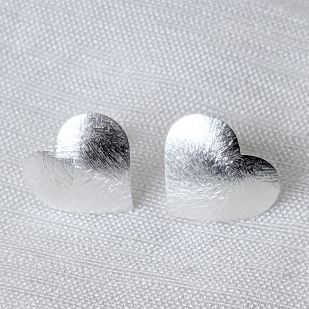 Nahaufnahme von zwei 925 Silber Ohrsteckern in Herzform mit markant gebürsteter, kratzmatter Oberfläche auf hellem Stoff.