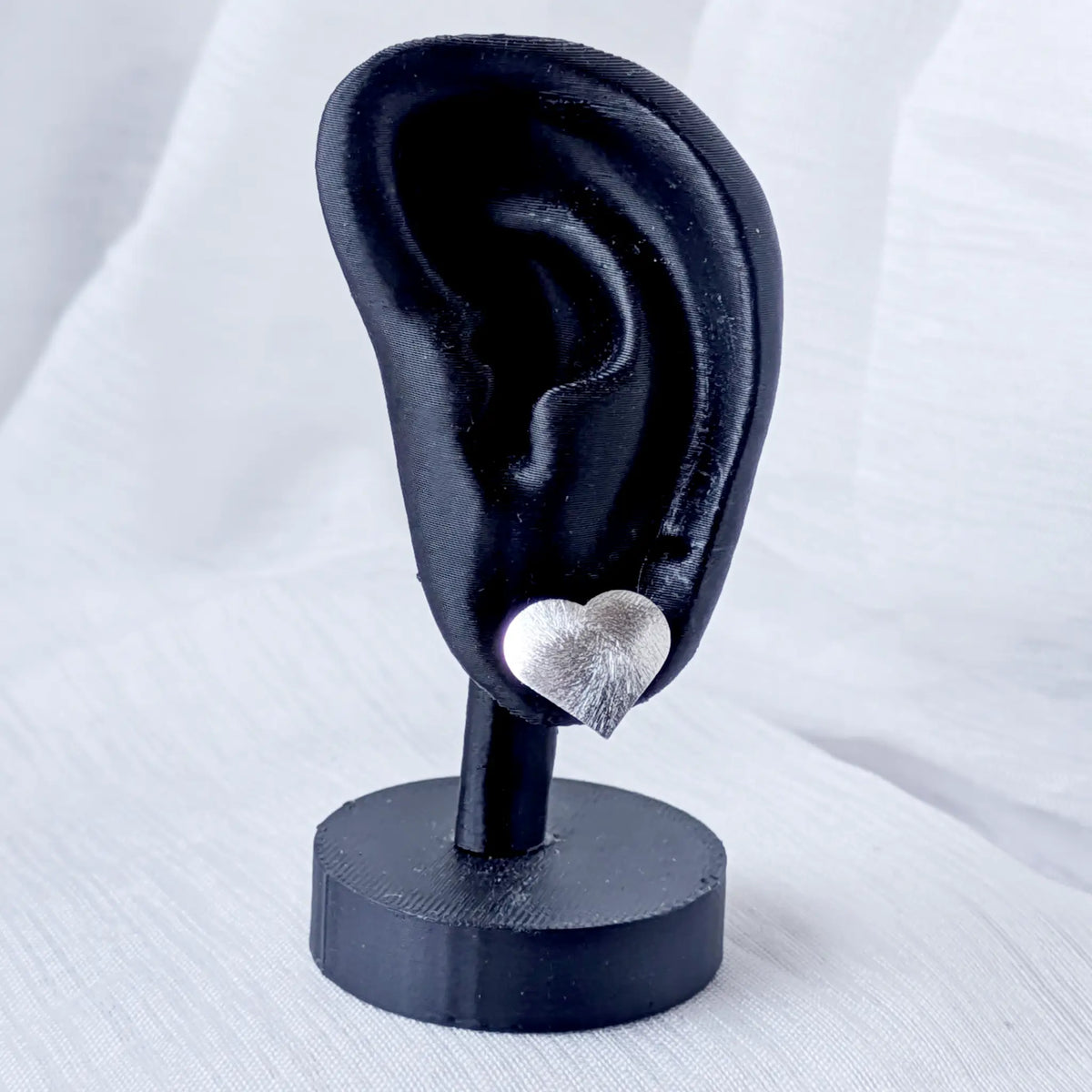 Ein strukturierter 925 Silber Herz-Ohrstecker präsentiert an einem schwarzen 3D-Ohr-Display, um die optimale Passform und Größe am Ohrläppchen zu verdeutlichen.