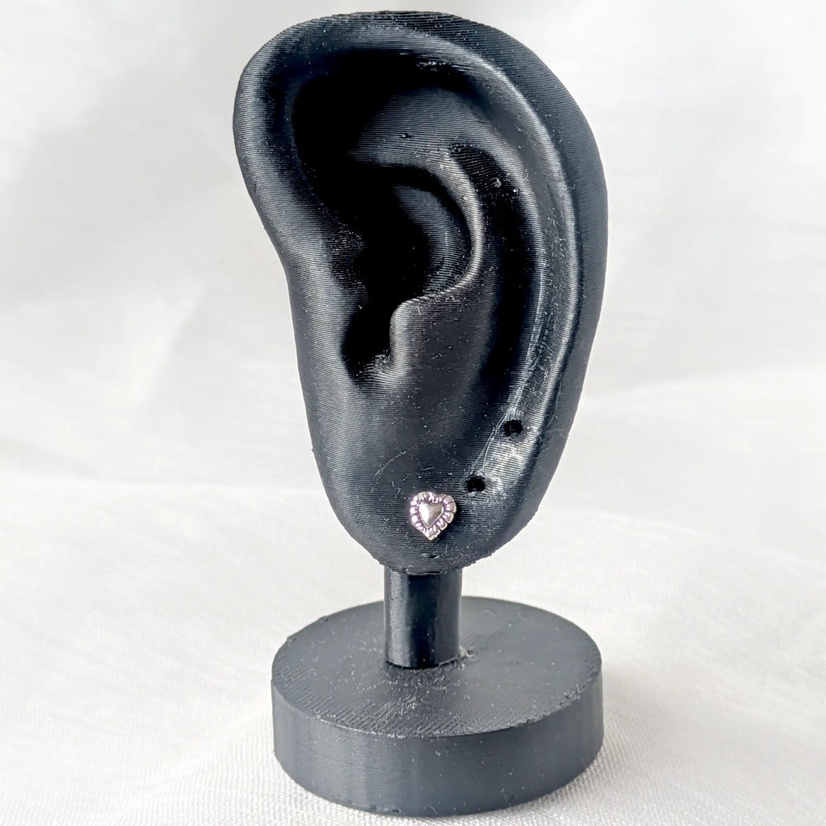 Ein zierlicher Herz-Ohrstecker aus Silber, präsentiert an einem schwarzen 3D-Ohr-Display, um die dezente Größe und Passform am Ohrläppchen zu veranschaulichen.