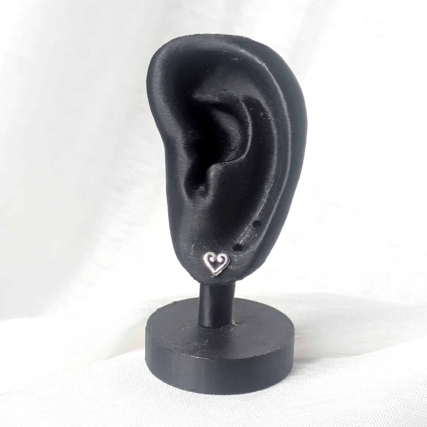 Ein kleiner silberner Herz-Ohrstecker präsentiert an einem schwarzen Ohr-Display zur Veranschaulichung der eleganten Passform am Ohrläppchen.