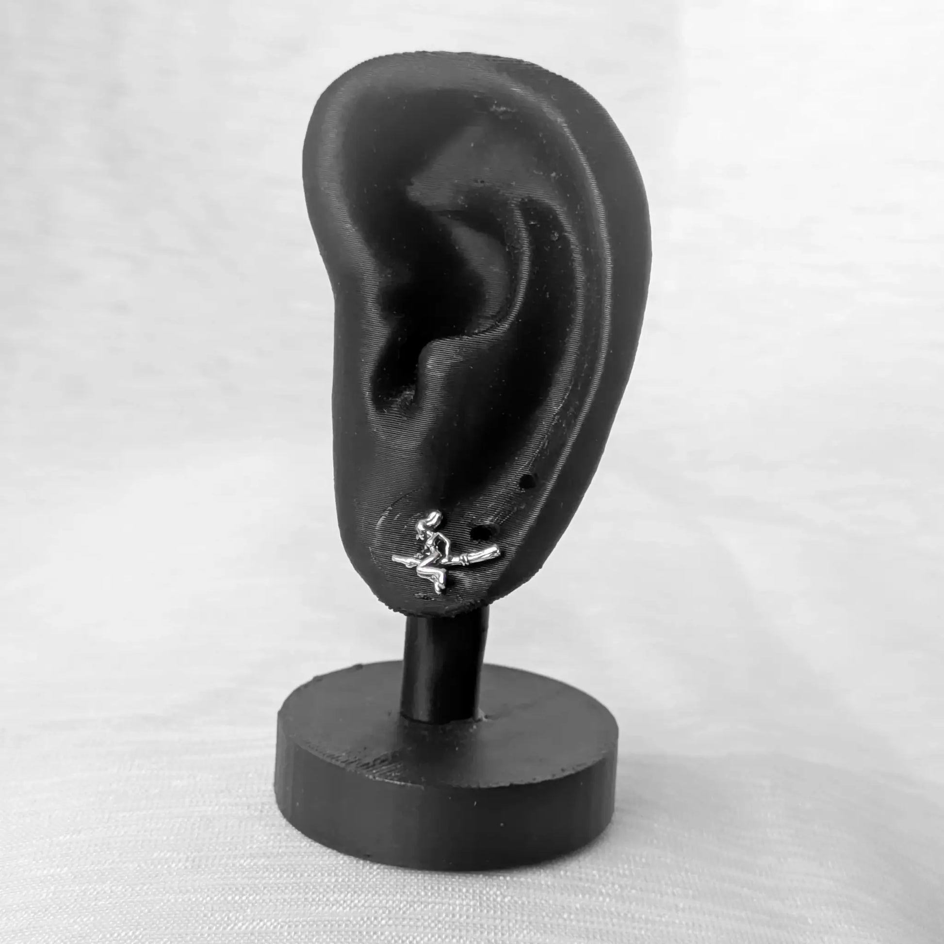 Ein silberner Ohrstecker in Form einer Hexe auf einem Besen, präsentiert an einem schwarzen Ohr-Modell.