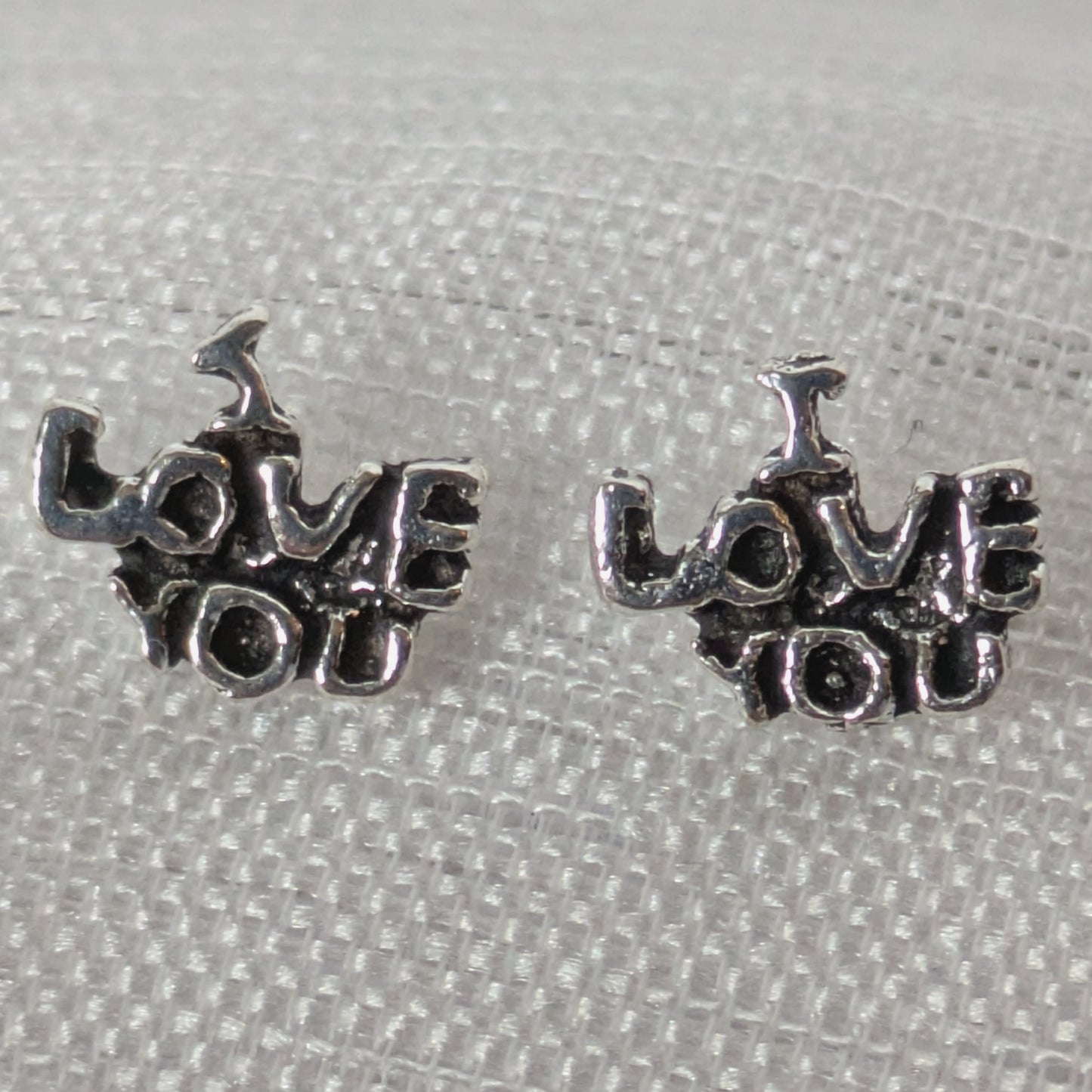 Frontalansicht der silbernen 'I Love You' Ohrstecker mit antikem Finish auf weißem strukturiertem Stoff.