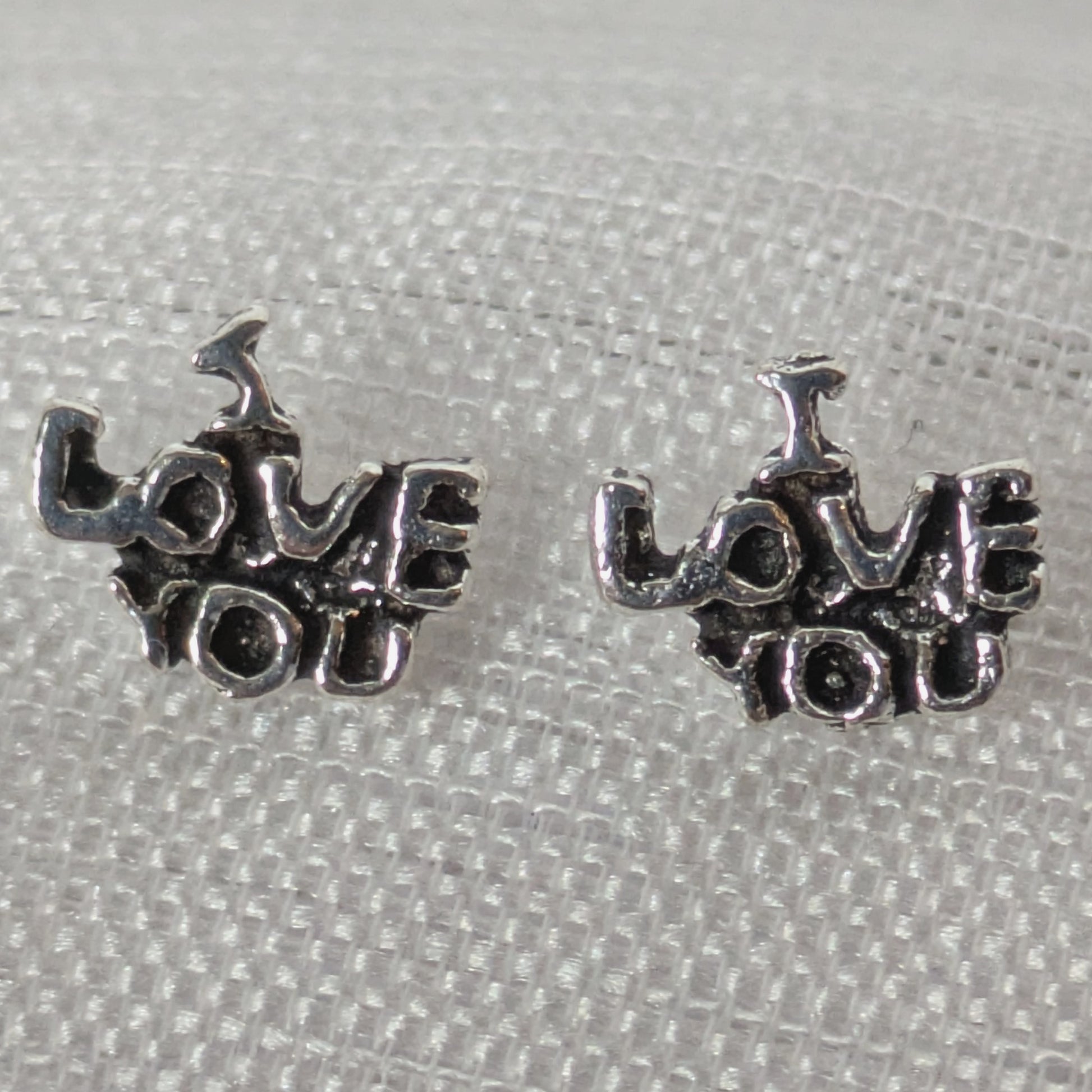 Frontalansicht der silbernen 'I Love You' Ohrstecker mit antikem Finish auf weißem strukturiertem Stoff.