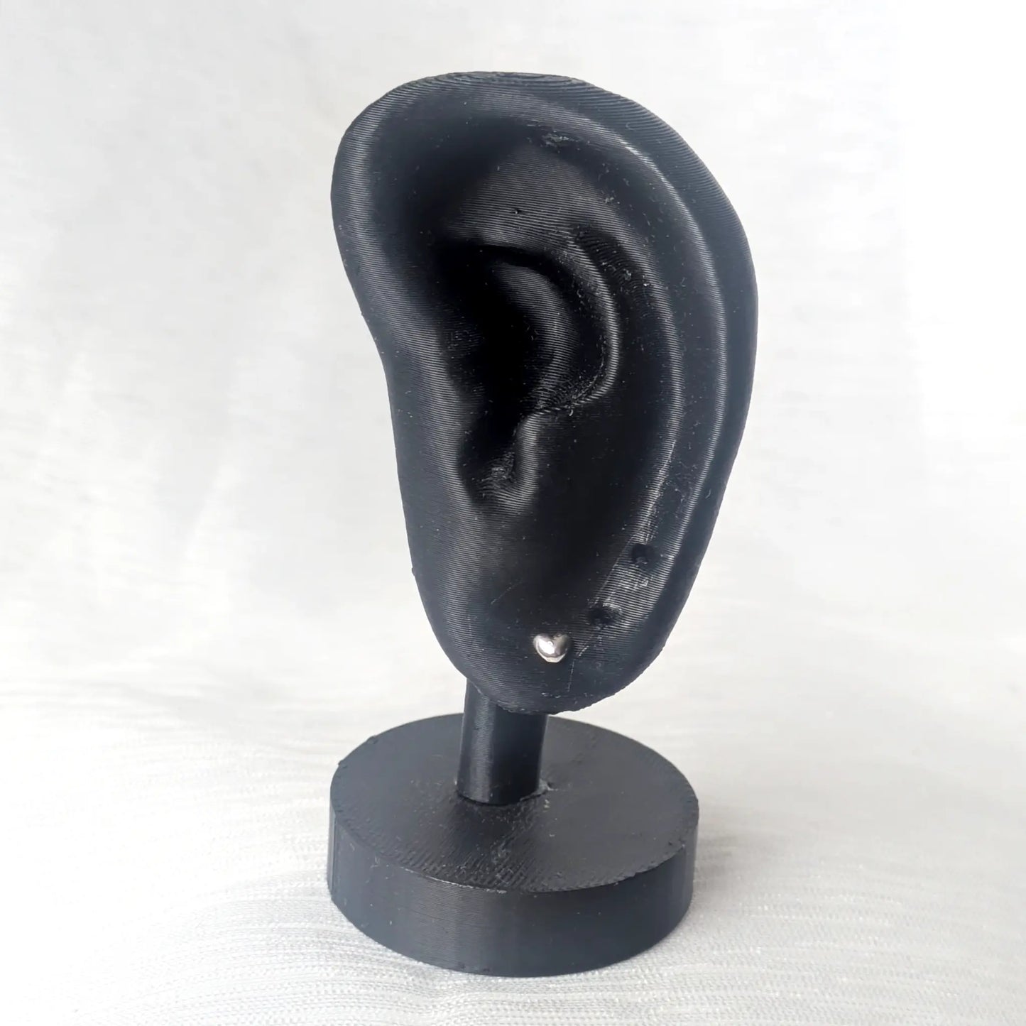Ein kleiner silberner Herz-Ohrstecker getragen an einem modernen, schwarzen Ohr-Display zur Veranschaulichung der grazilen Proportion am Ohr.