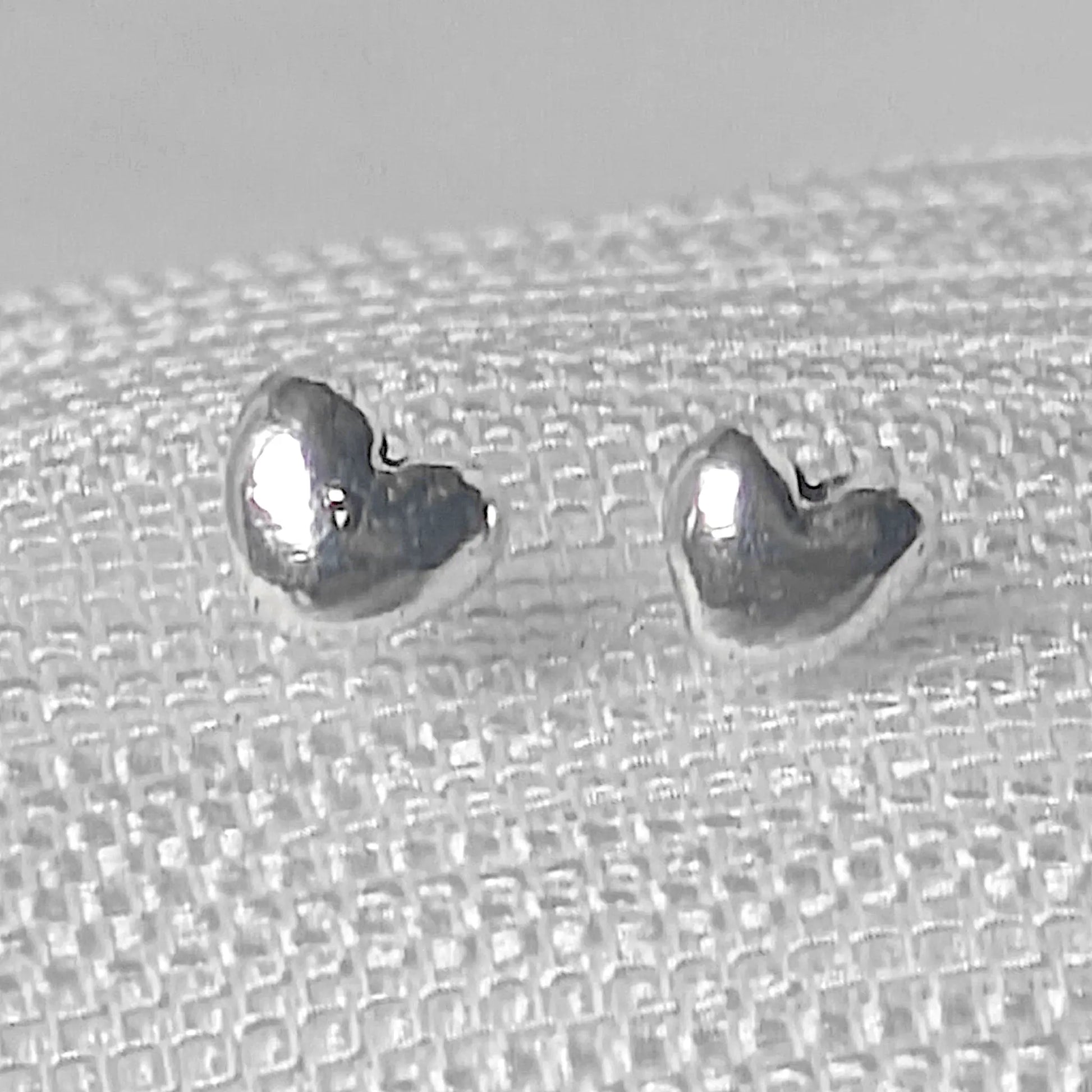 Nahaufnahme von zwei glänzenden, dreidimensionalen Herz-Ohrsteckern aus echtem 925 Sterling Silber auf einer hellen Texturstruktur