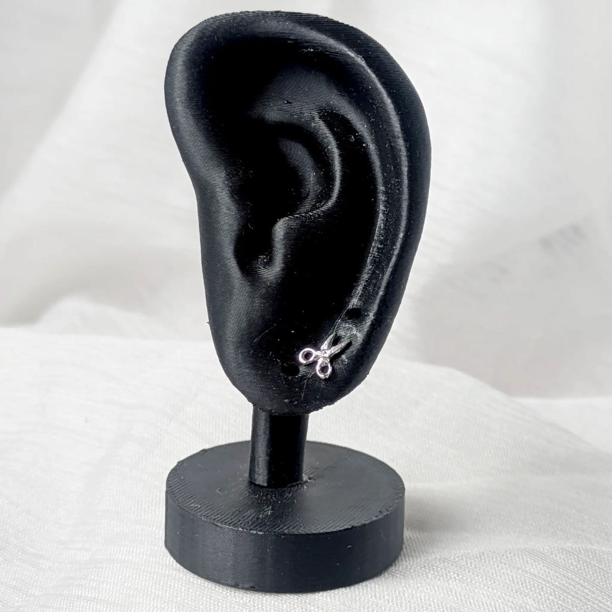 Ein kleiner, silberner Scheren-Ohrstecker, dezent getragen am Ohrläppchen eines schwarzen 3D-Modell-Ohrs zur Größenveranschaulichung.