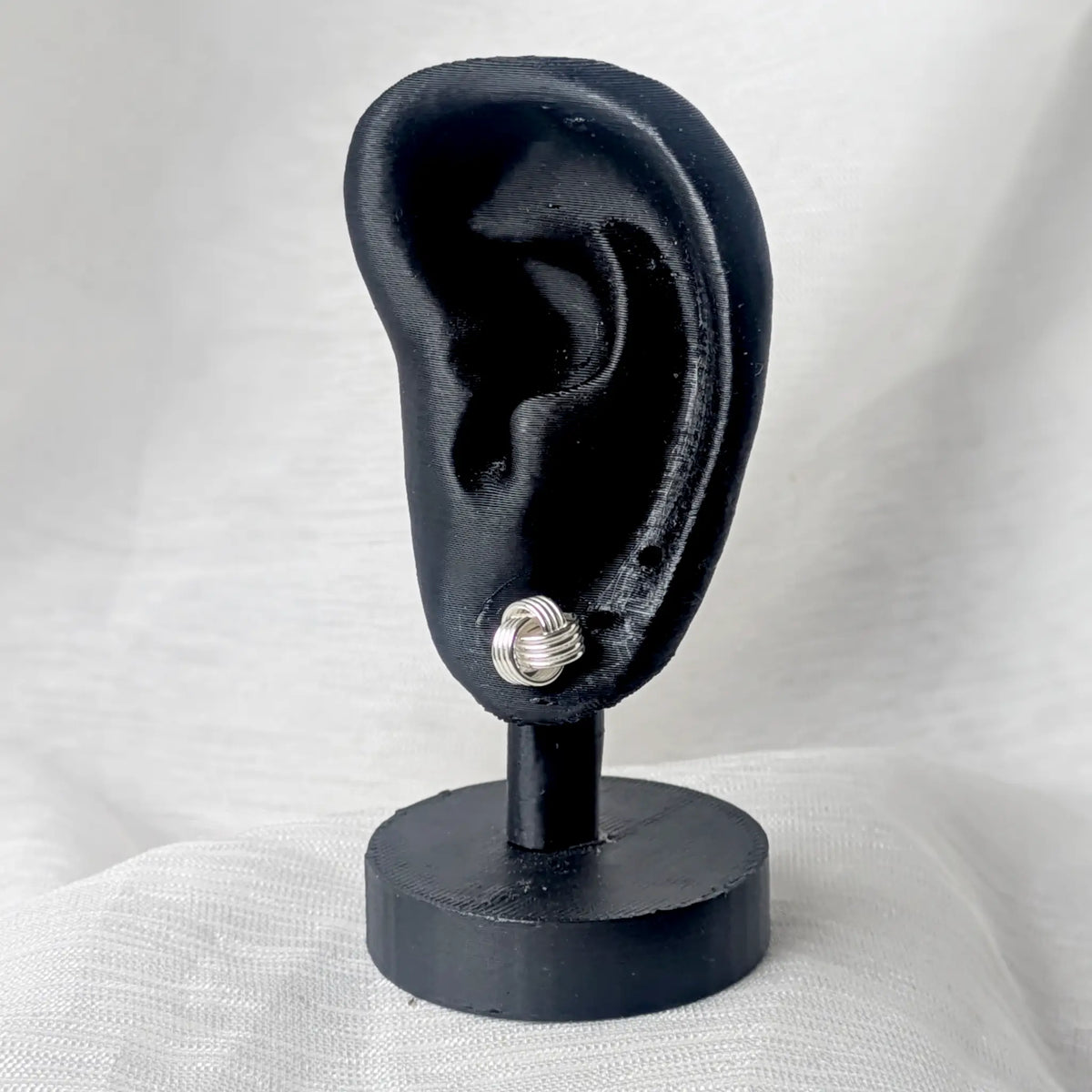 Glänzender Silber-Ohrstecker im Knoten-Design, ideal positioniert am Ohrläppchen eines schwarzen Modell-Ohrs zur Demonstration der Proportionen.