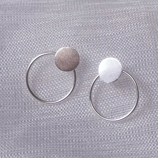 Nahaufnahme von zwei geometrischen 925 Silber Ohrsteckern mit rundem Plättchen und filigranem Kreis auf hellem Stoffhintergrund.