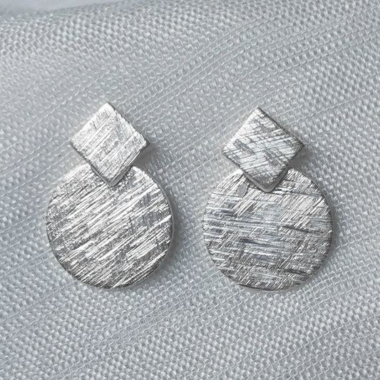 Nahaufnahme von geometrischen 925 Silber-Ohrsteckern mit detailreich gebürsteter Oberfläche in mattierter Eiskratz-Optik.