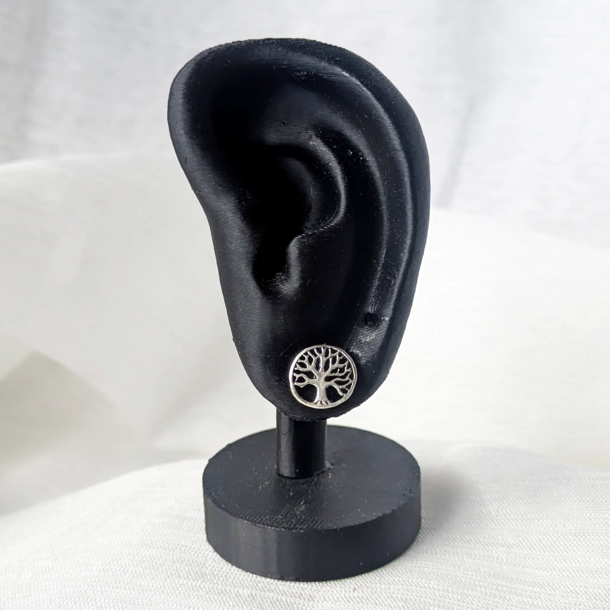 Ein runder Lebensbaum-Ohrstecker aus Silber, platziert an einem schwarzen, 3D-gedruckten Ohrläppchen-Display.