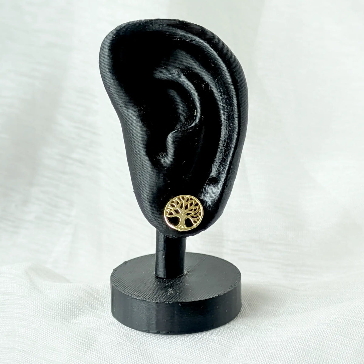 Ein runder, vergoldeter Lebensbaum-Ohrstecker, elegant getragen an einem schwarzen Ohr-Display zur Veranschaulichung der dezenten Proportionen am Ohr.
