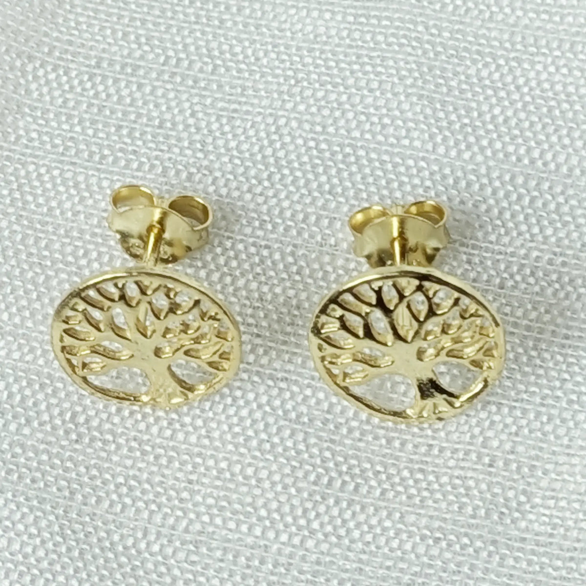 Frontalansicht der glänzenden, goldenen Baum-des-Lebens Ohrringe, liegend auf einem hellen, leicht strukturierten Stoff.