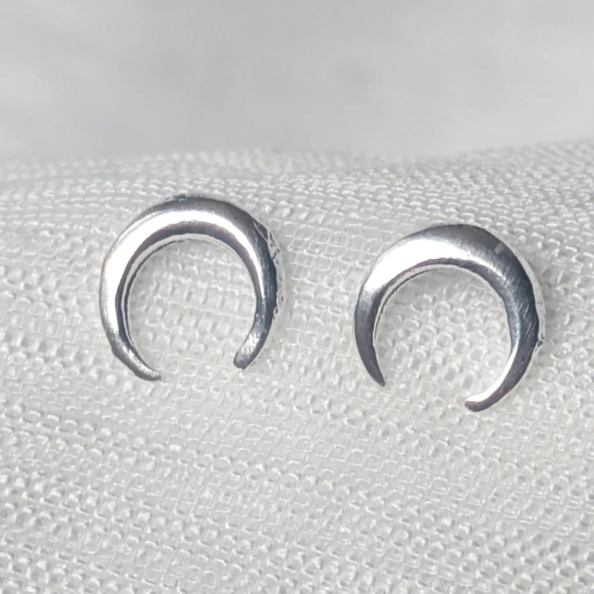 Nahaufnahme von zwei glänzenden, nach unten geöffneten Halbmond-Ohrsteckern aus 925 Sterling Silber auf hellem Stoff.