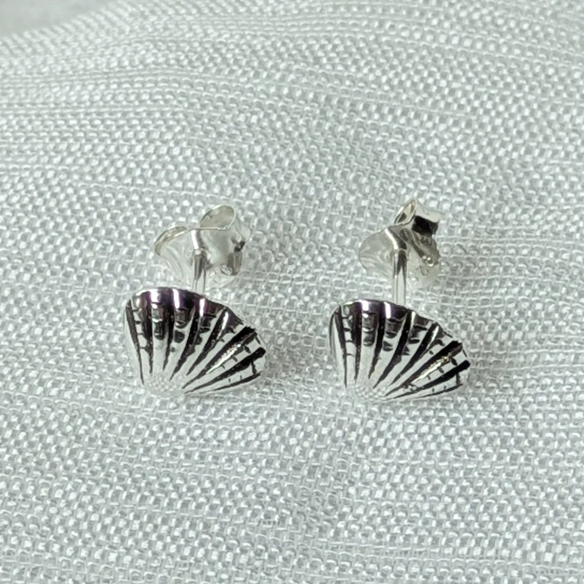 Frontalansicht der kleinen silbernen Ohrstecker in Muschelform, liegend auf einem hellen, strukturierten Stoff.