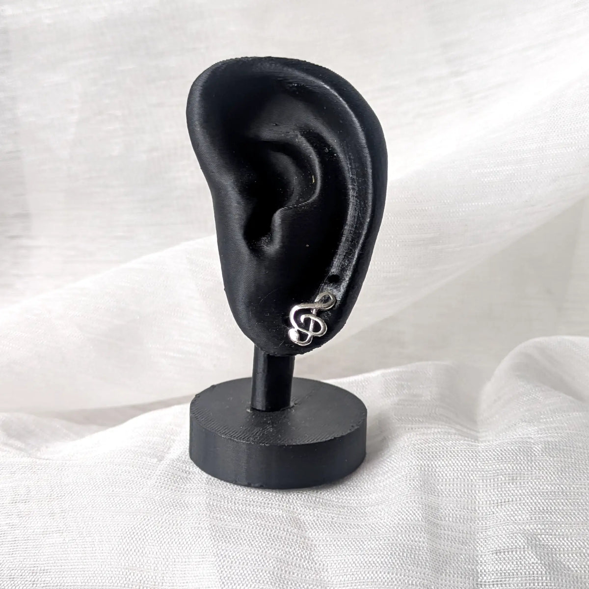 Ein silberner Notenschlüssel-Ohrstecker, präsentiert an einem schwarzen Ohr-Display zur Veranschaulichung der dezenten Proportionen am Ohrläppchen.
