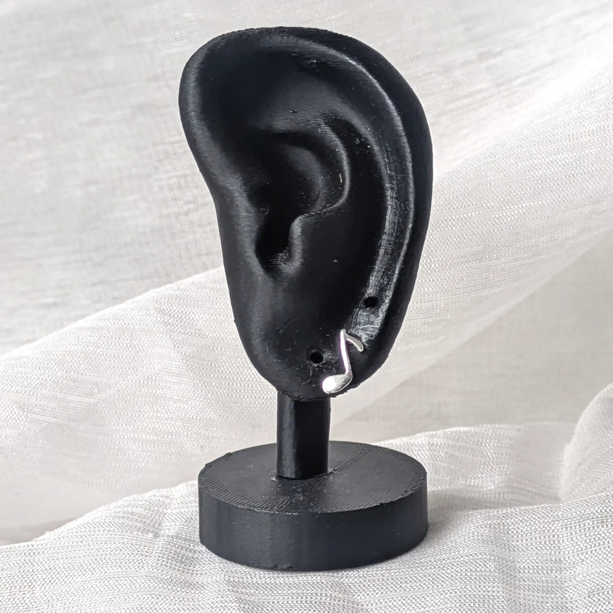 Ein silberner Notenschlüssel-Ohrstecker, präsentiert an einem schwarzen 3D-Modell-Ohr zur Veranschaulichung der dezenten Proportionen am Ohrläppchen.