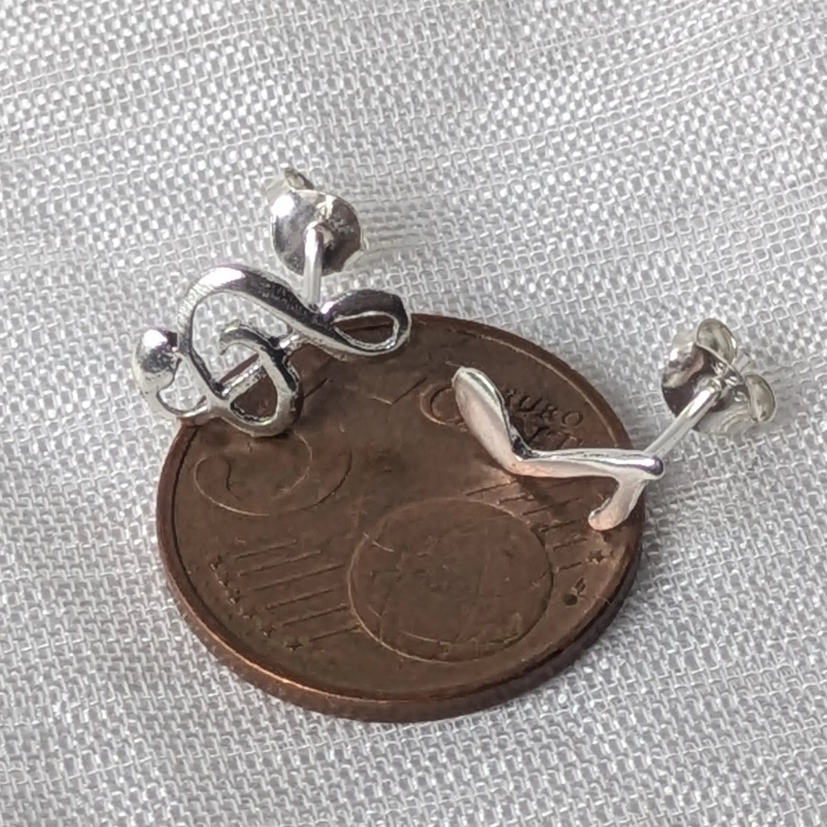 Größenvergleich der kleinen asymmetrischen Silberohrstecker (Notenschlüssel und Note), flach liegend auf einer 5-Cent-Münze.