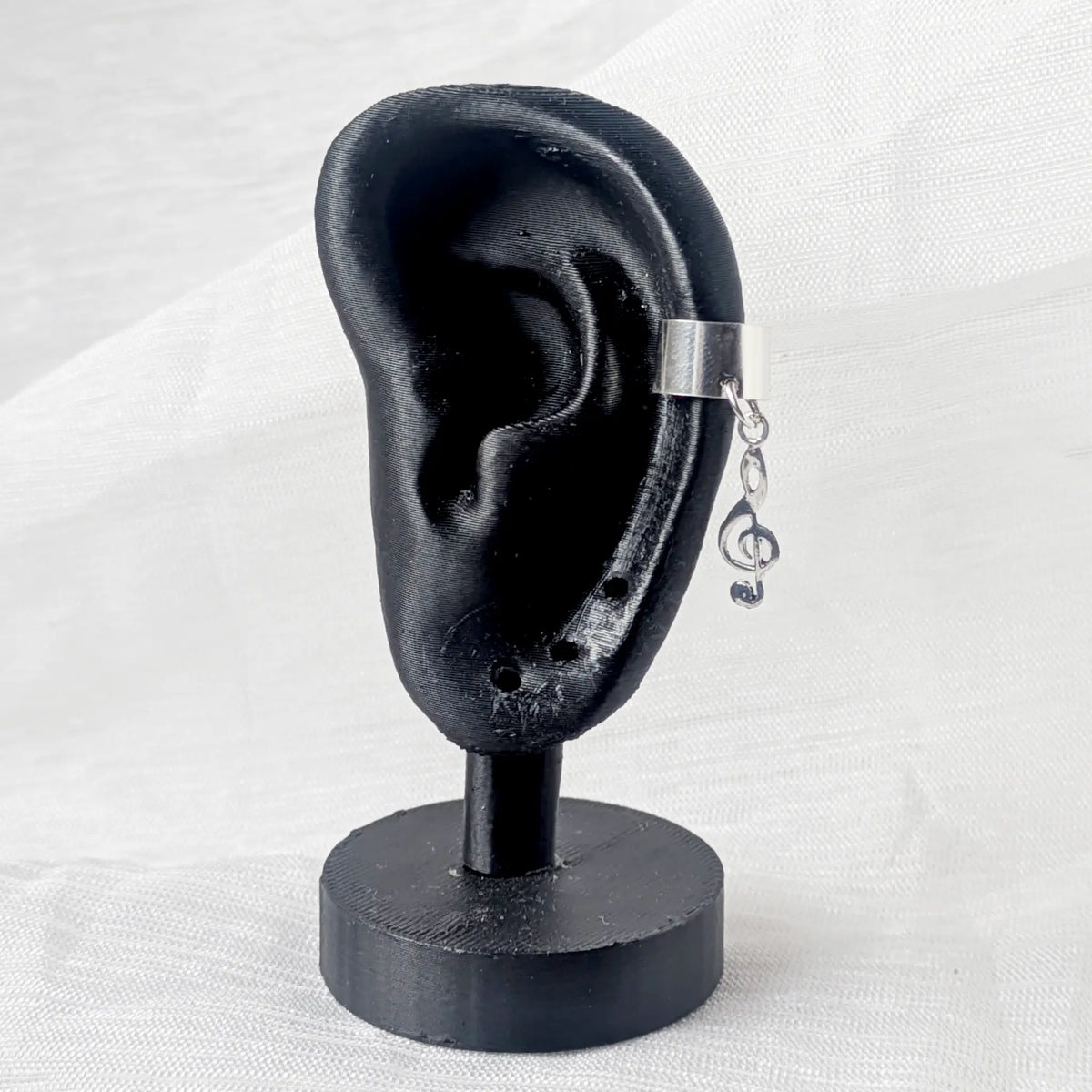 Silberner Earcuff mit Notenschlüssel getragen an der seitlichen Ohrmuschel eines schwarzen Ohr-Displays.

