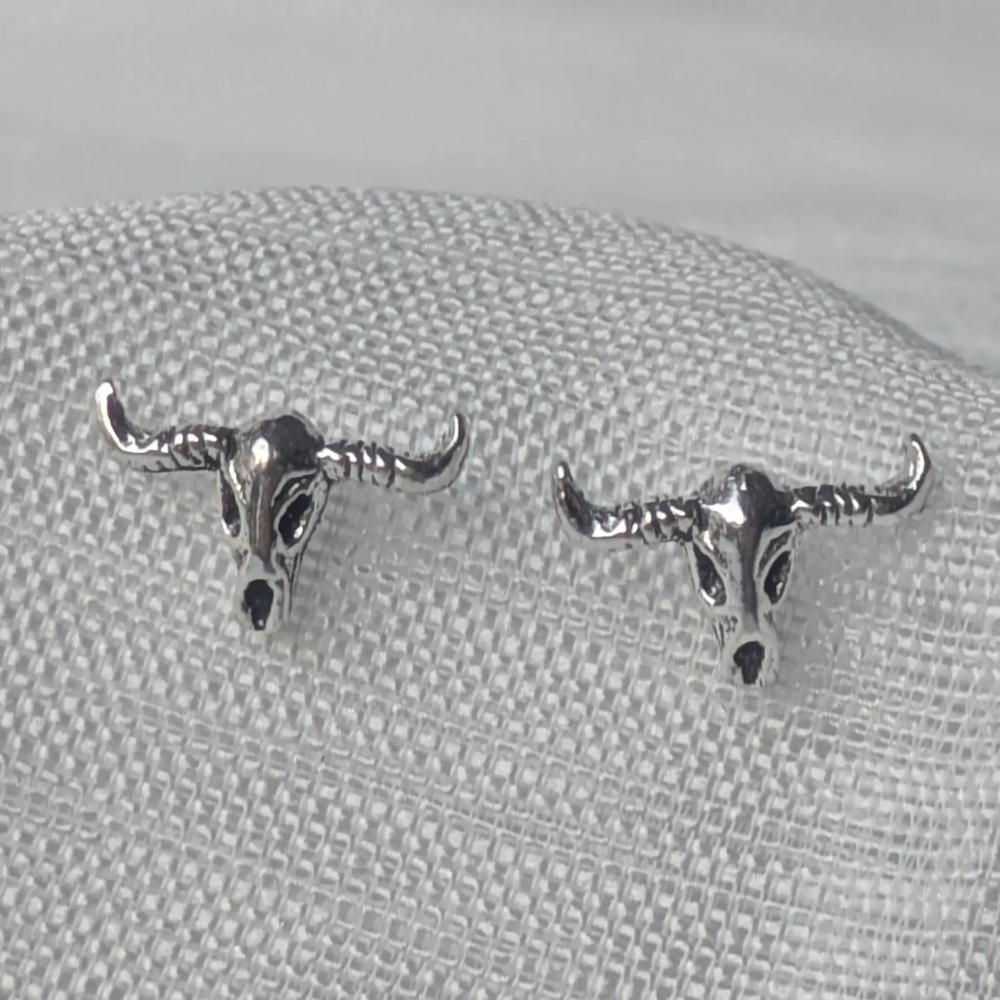 Nahaufnahme von zwei detaillierten Büffelschädel-Ohrsteckern aus oxidiertem 925 Sterling Silber auf weißem Stoffhintergrund.