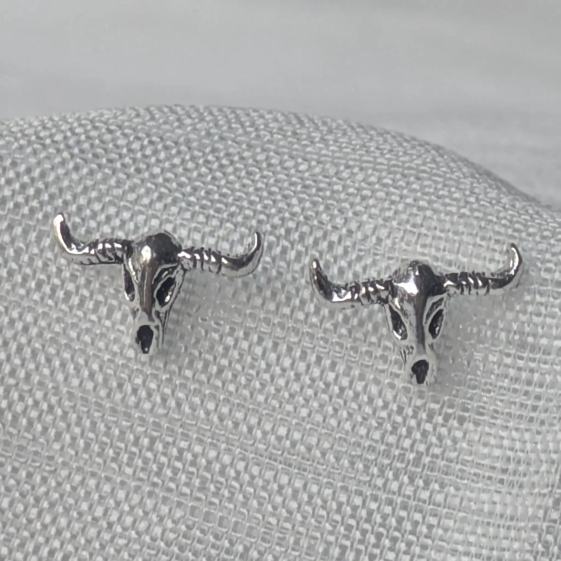 Nahaufnahme von zwei detaillierten Büffelschädel-Ohrsteckern aus oxidiertem 925 Sterling Silber auf weißem Stoffhintergrund.