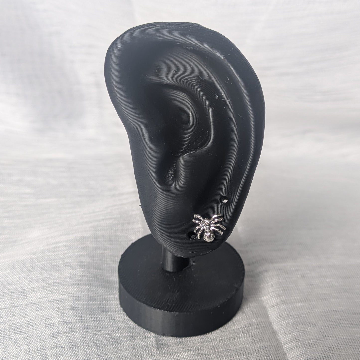 Kleine Spinnen‑Ohrringe aus Sterling Silber, glänzende Oberfläche auf schwarzem Ohr.