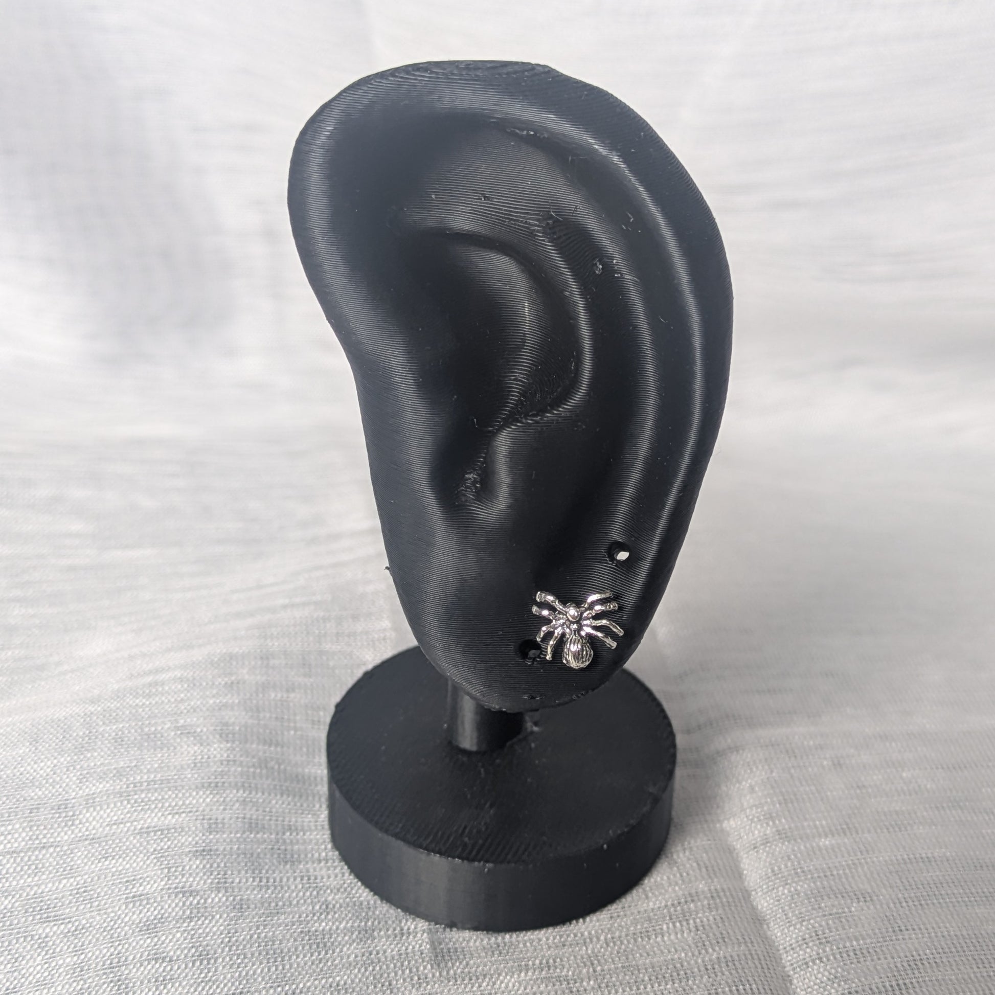 Kleine Spinnen‑Ohrringe aus Sterling Silber, glänzende Oberfläche auf schwarzem Ohr.