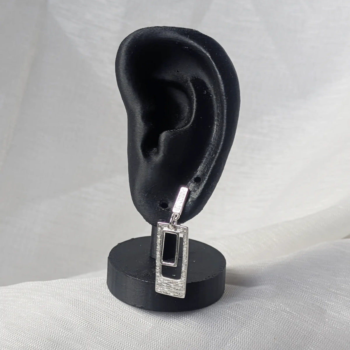 Hängender, rechteckiger Silber-Ohrstecker getragen an einem schwarzen Ohr-Display zur Veranschaulichung der Proportionen am Ohrloch.
