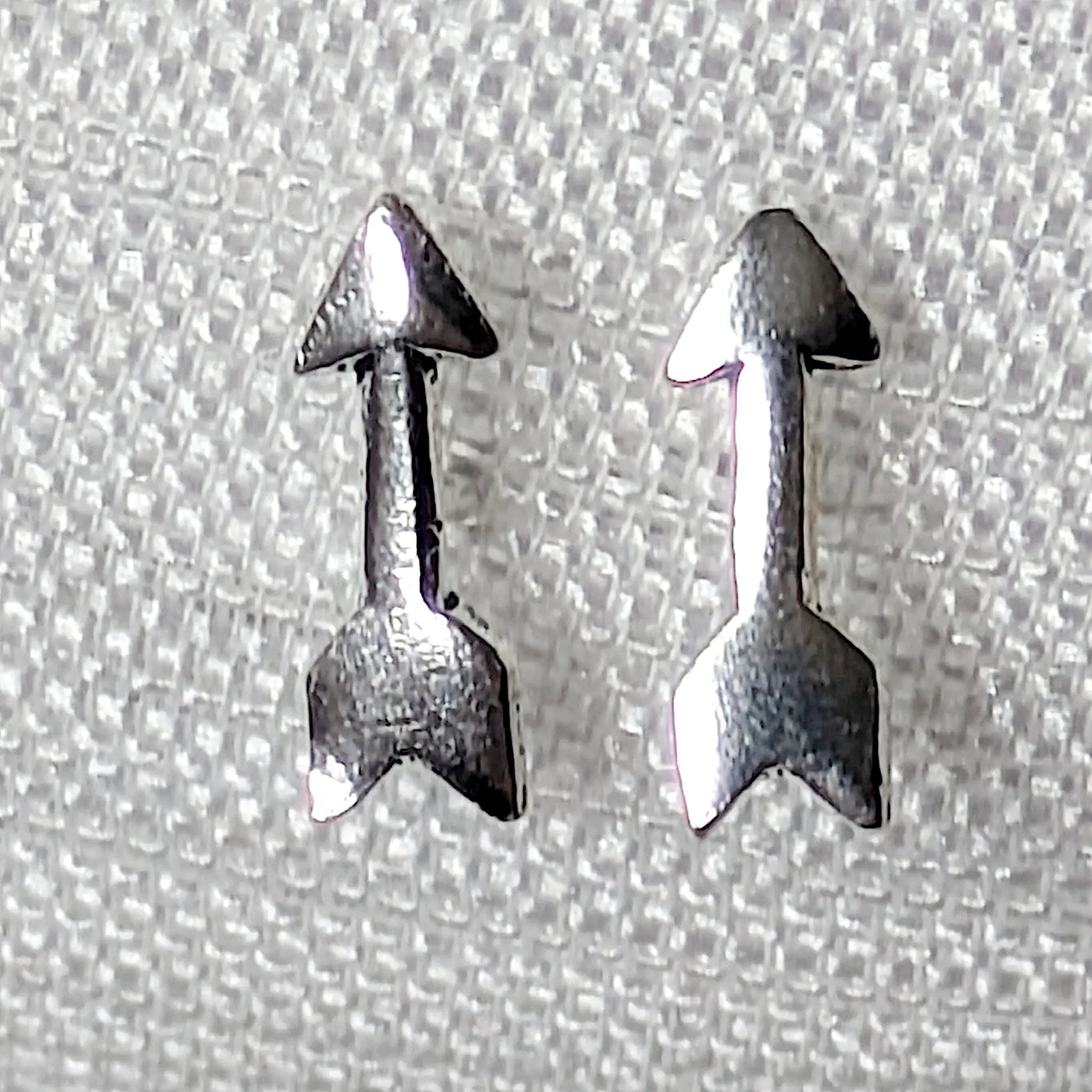 Nahaufnahme von zwei kleinen Pfeil-Ohrsteckern aus glänzendem 925 Silber auf hellem Stoffhintergrund.